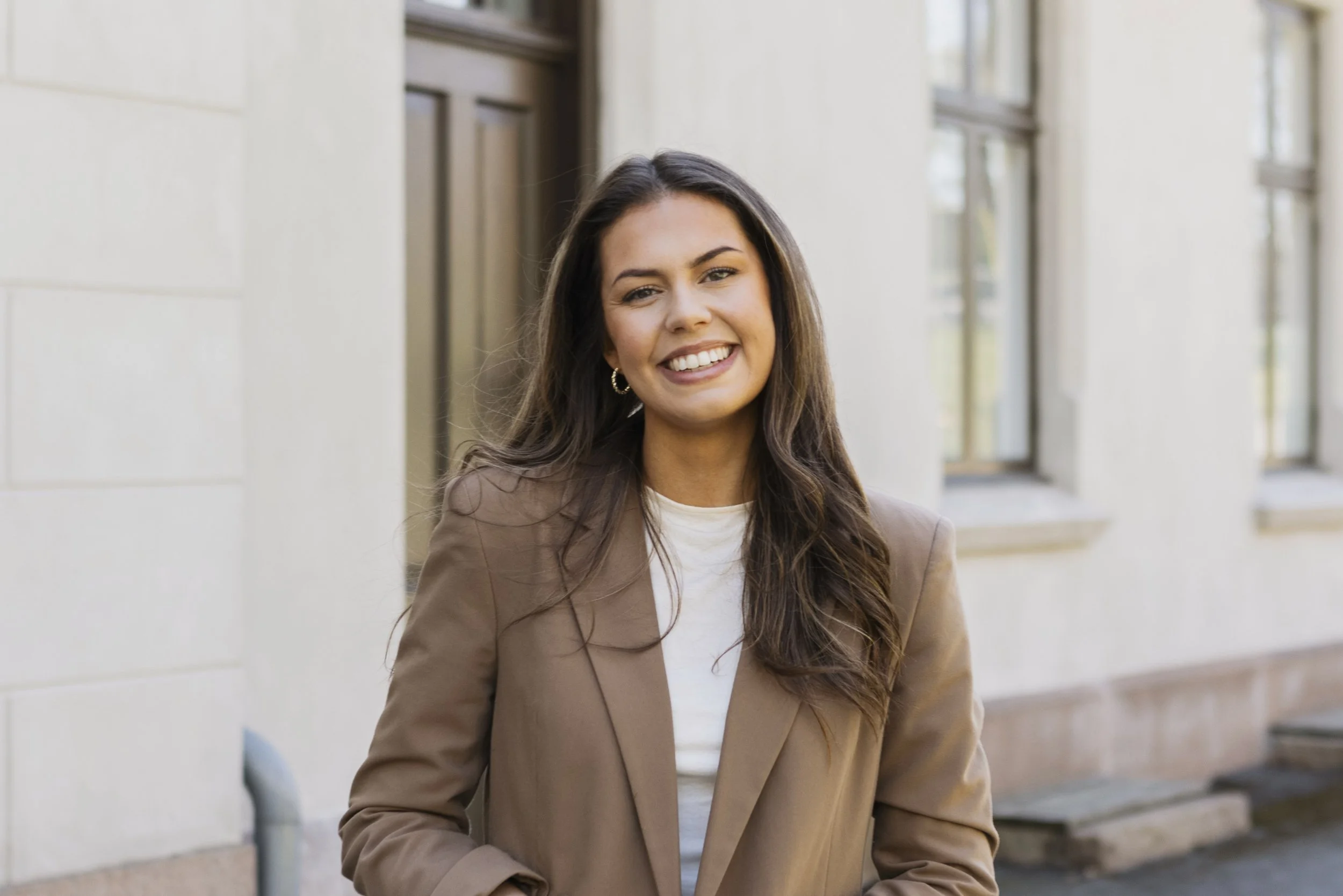 A woman with long brown hair wearing a beige blazer and white top, smiling outdoors in front of a building with windows and a white wall.