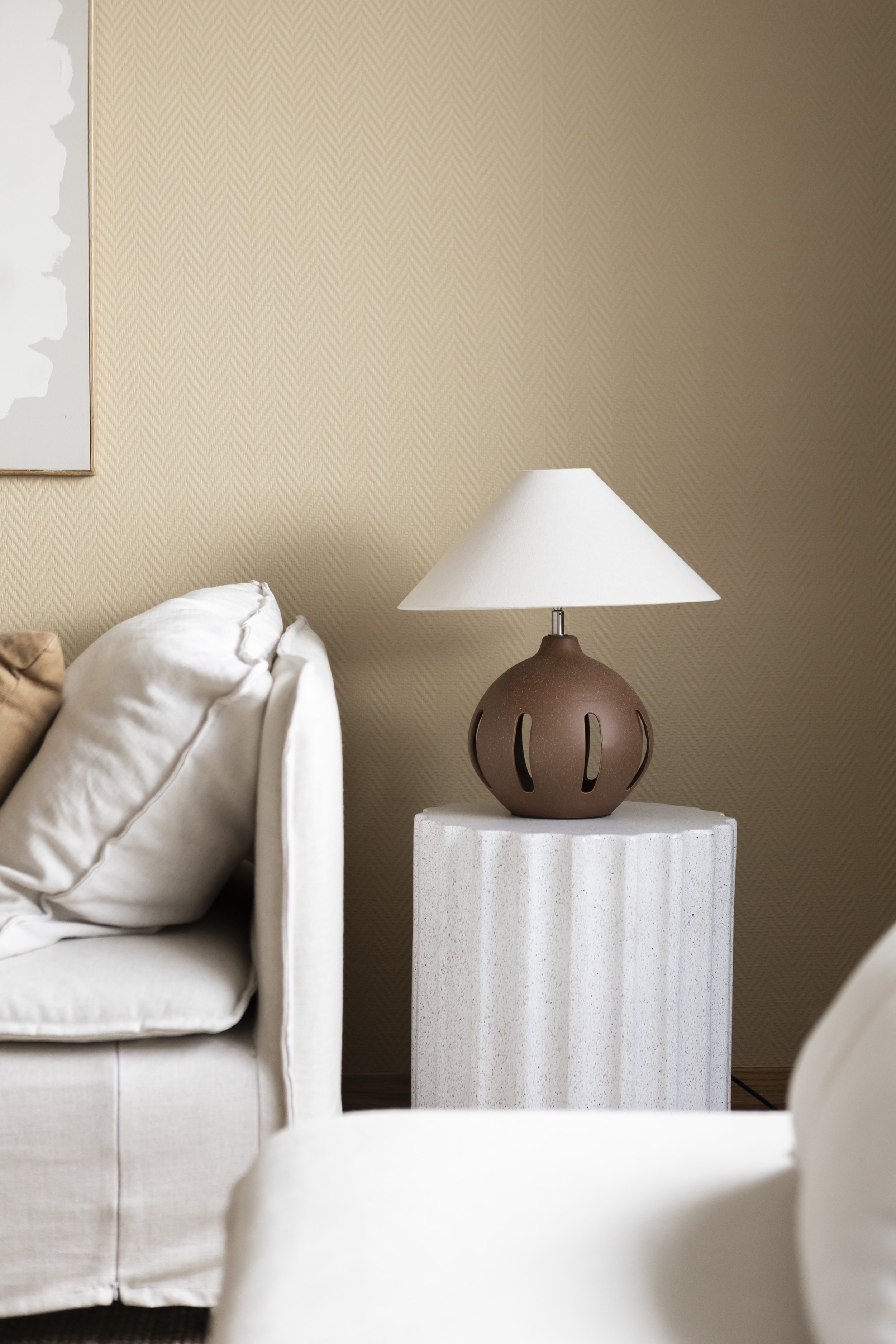 Living room corner with cream sofa, white pillow, textured wallpaper, beige table lamp with brown base, and a textured white side table.