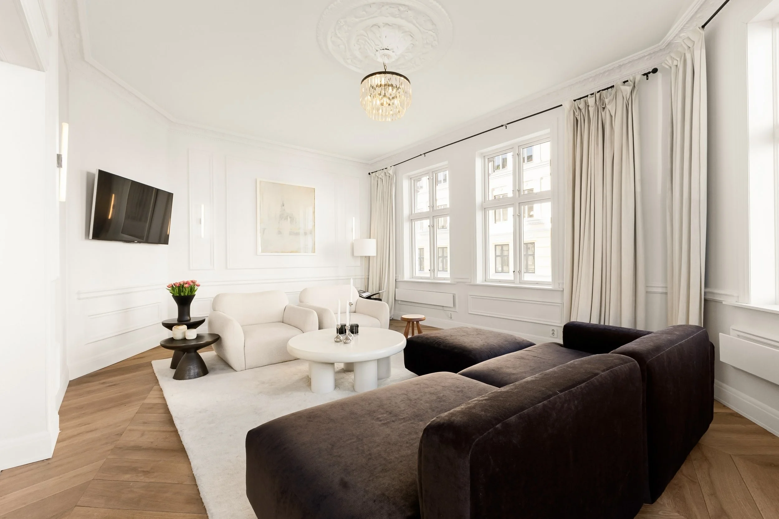 Bright living room with white walls and large windows, featuring a dark brown sofa, white armchair, glass coffee table with decor, a flower vase, wall-mounted TV, and a ceiling light fixture.