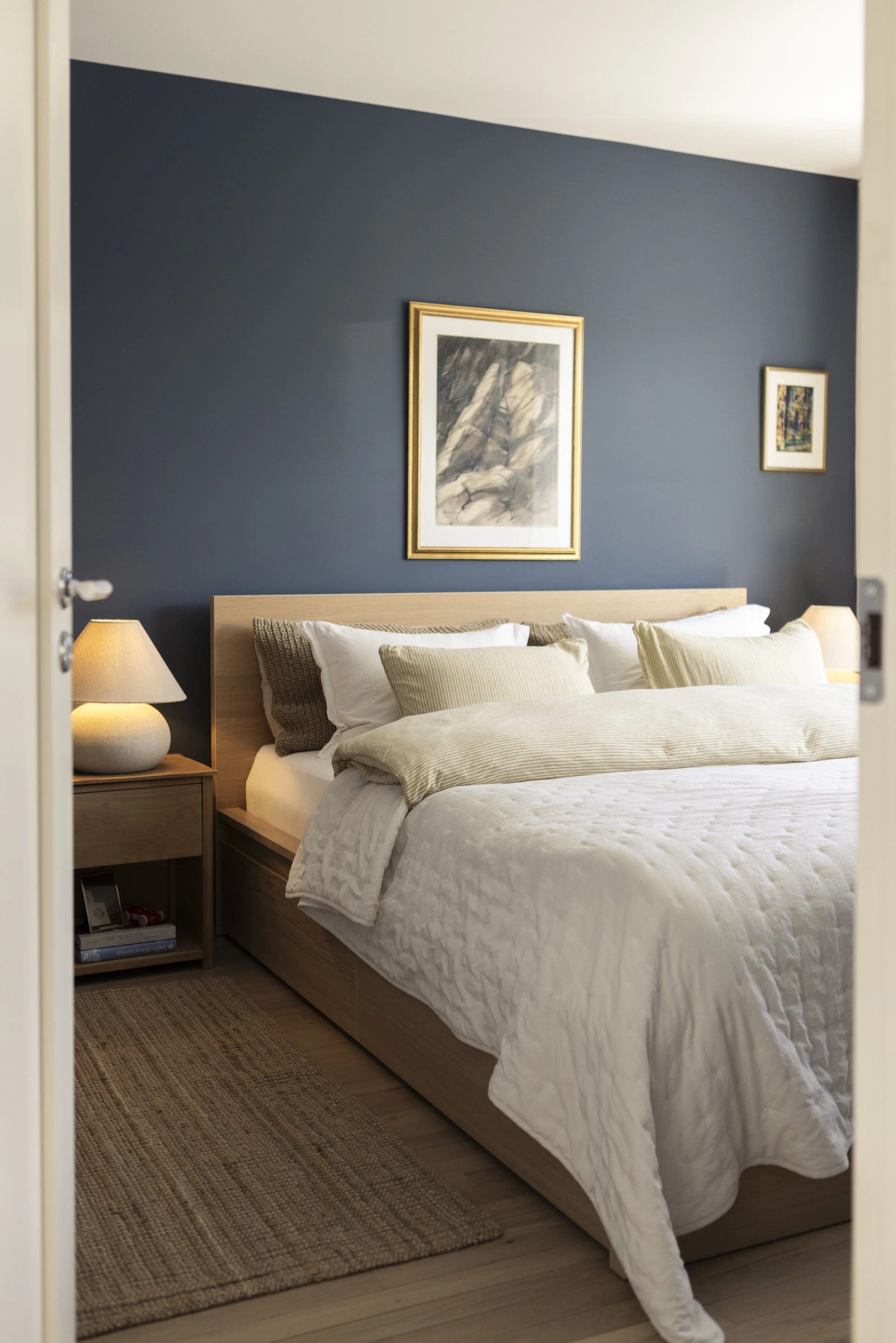 A neatly made bed with a light wood frame, topped with white sheets and various beige and white pillows. On the nightstand, there is a white table lamp and a few books underneath. The wall behind the bed is painted dark blue, decorated with two framed art pictures.