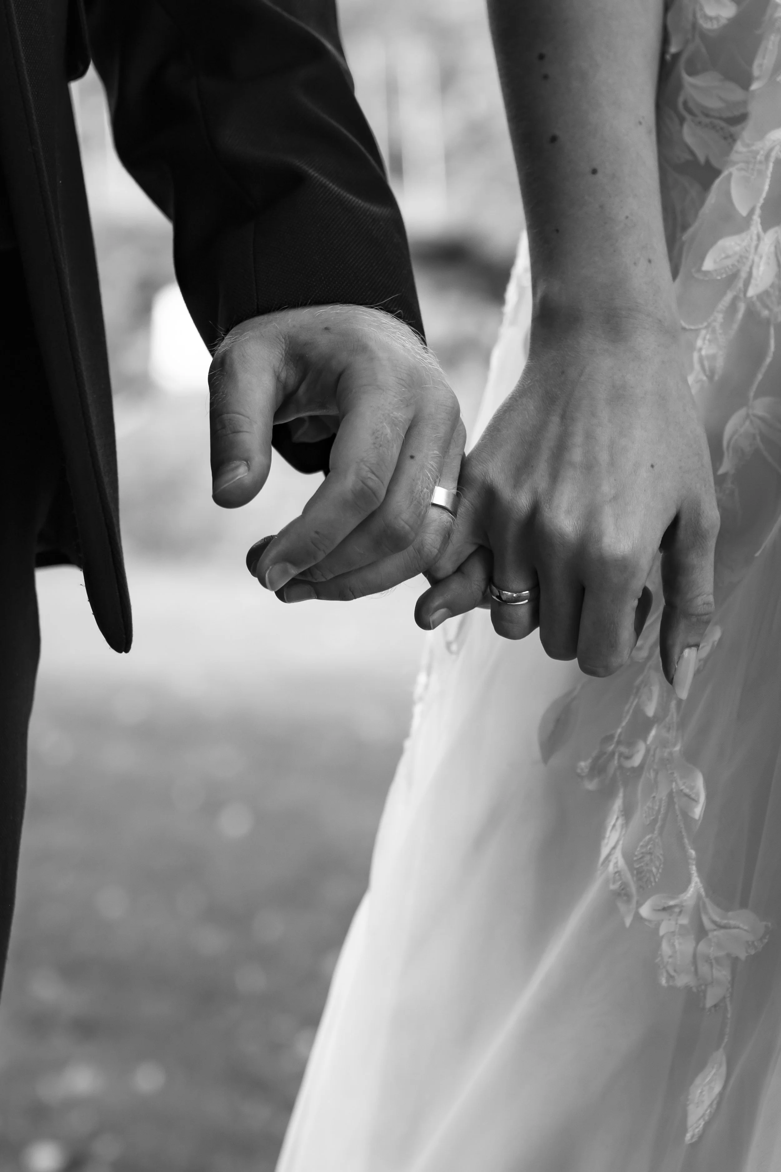 Close-up black and white photograph of a man and woman holding hands, with wedding rings visible on their ring fingers, dressed in formal attire, suggesting a wedding or romantic moment.