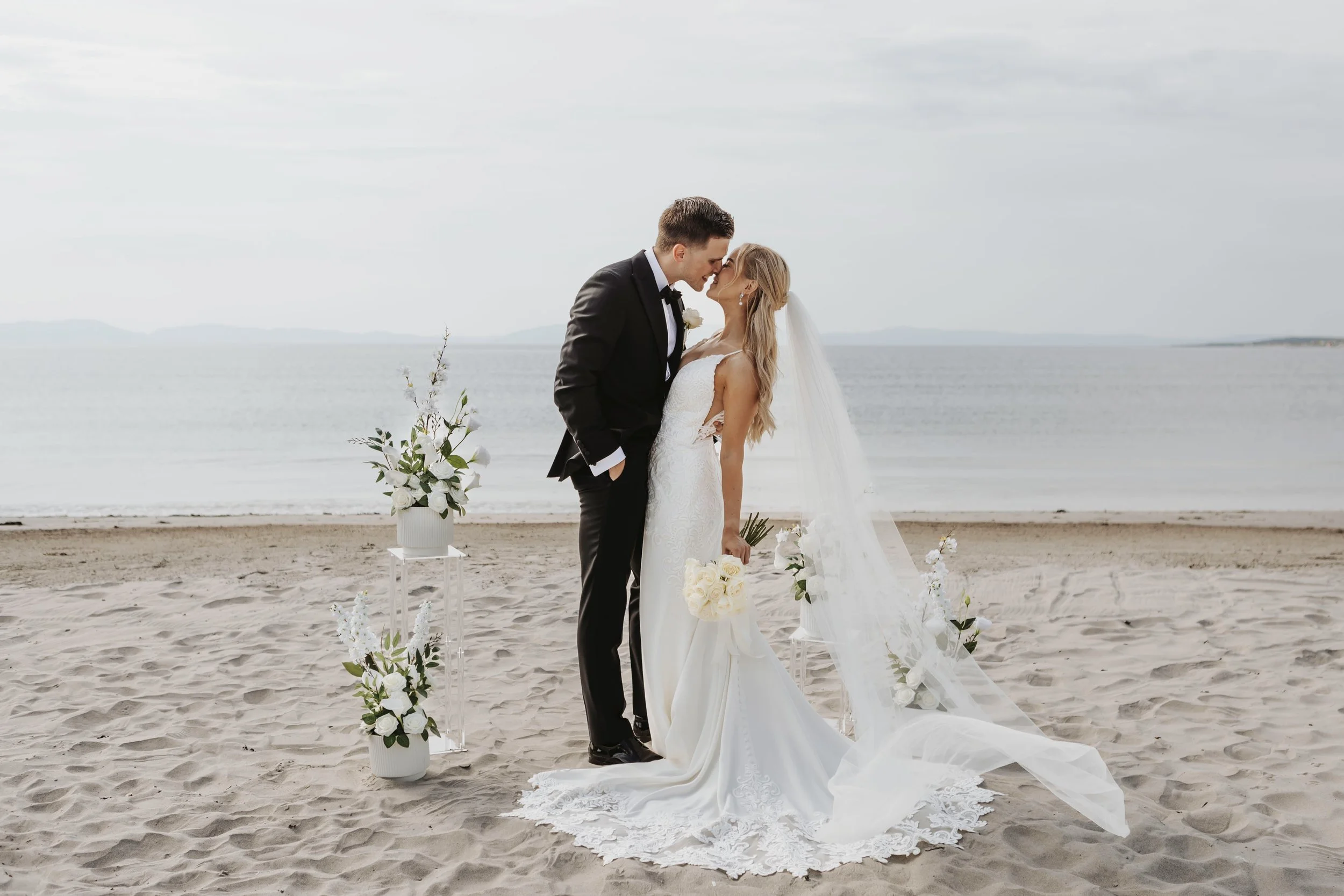 A bride and groom share an intimate moment during their wedding on the beach with the ocean in the background. The bride is holding a bouquet of white roses, and the groom is dressed in a black tuxedo. Decorated with white flowers and greenery, the ceremony setup is simple yet elegant.