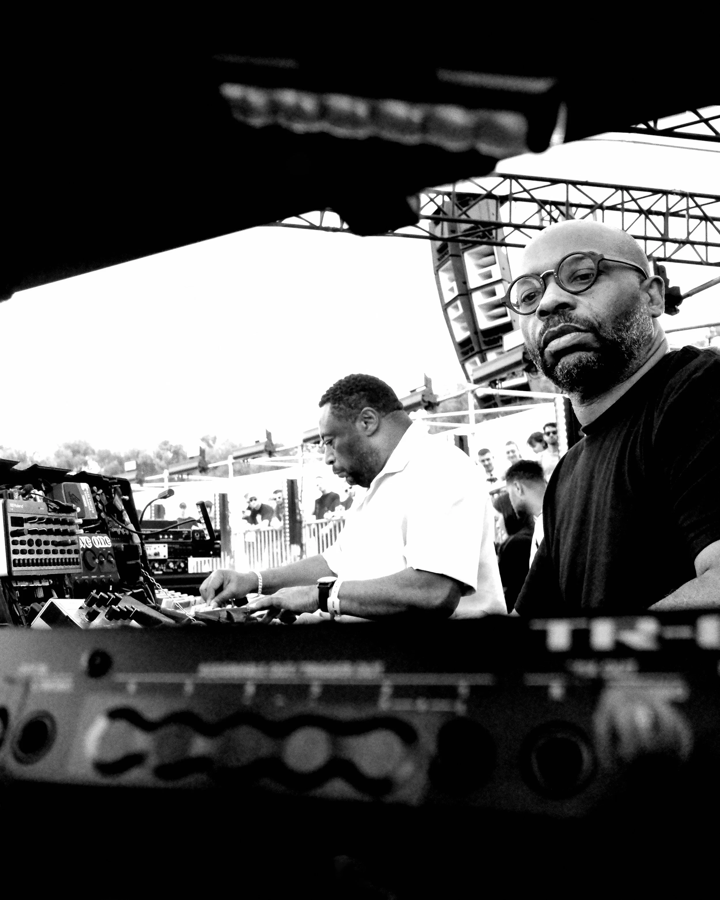 A black and white photo of two men at an outdoor event, one with glasses and a beard looking directly at the camera, the other with glasses focusing on equipment, with a crowd and metal structures in the background.