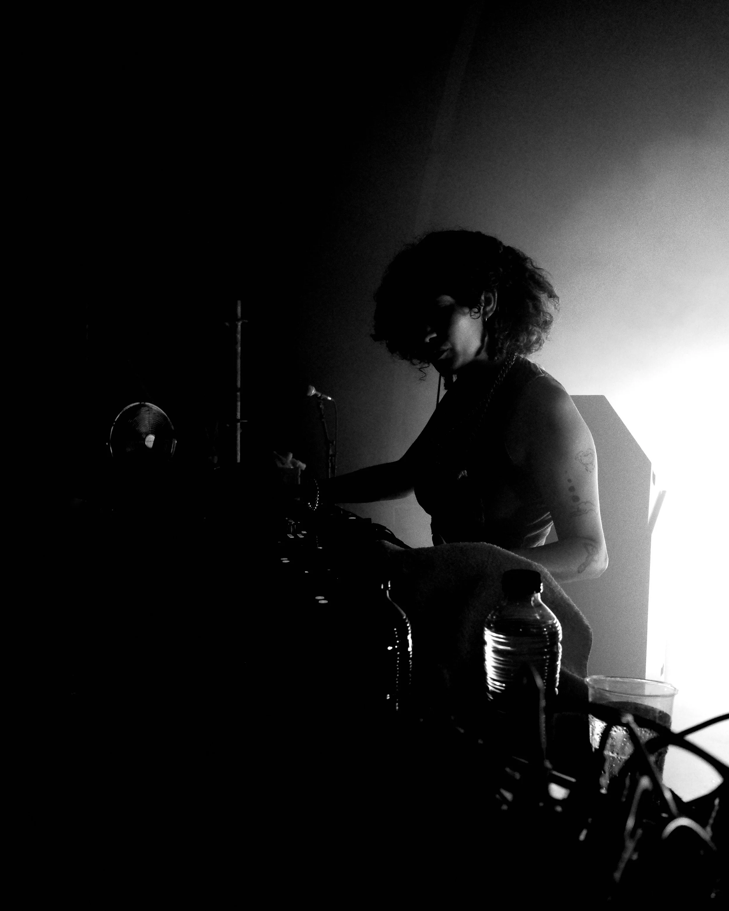 Black and white photo of a woman with curly hair, Mia Koden , operating DJ equipment in a dimly lit room with a bright light behind her at Dekmantel Festiva. Various objects including bottles and a fan are visible in the foreground.