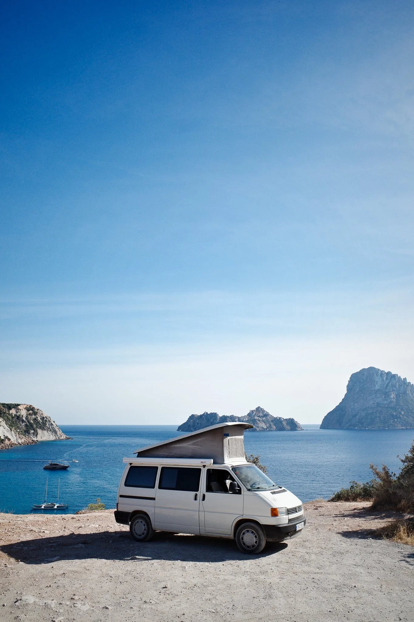 A white camper van parked on a dirt area near the coast, with mountains, a blue sea, and a boat in the background under a clear sky.