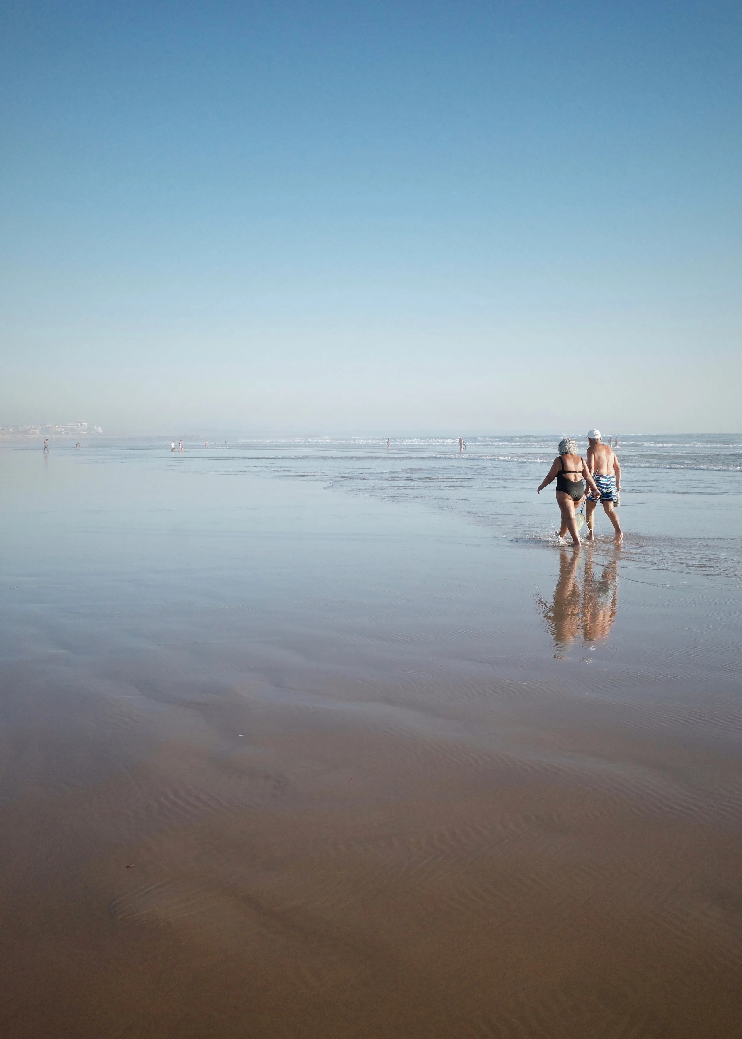 A beach scene with two elderly people walking hand in hand along the shoreline, with other beachgoers and buildings visible in the distance under a clear blue sky.