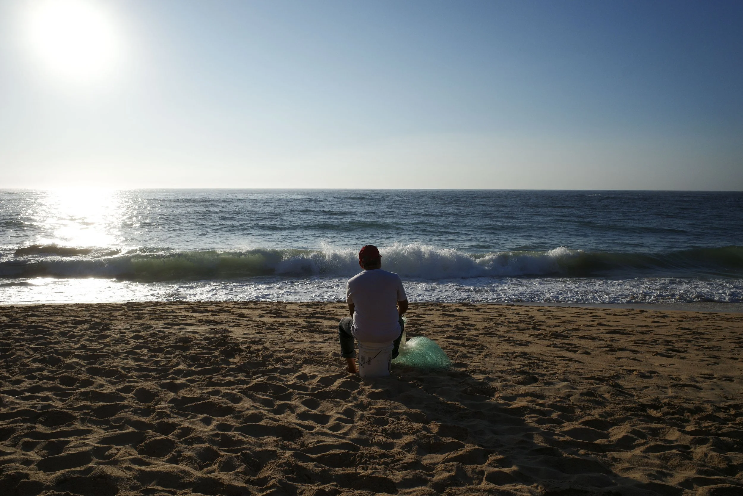 A person sitting on a sandy beach facing the ocean, with waves breaking near the shore and the sun shining brightly in the sky.