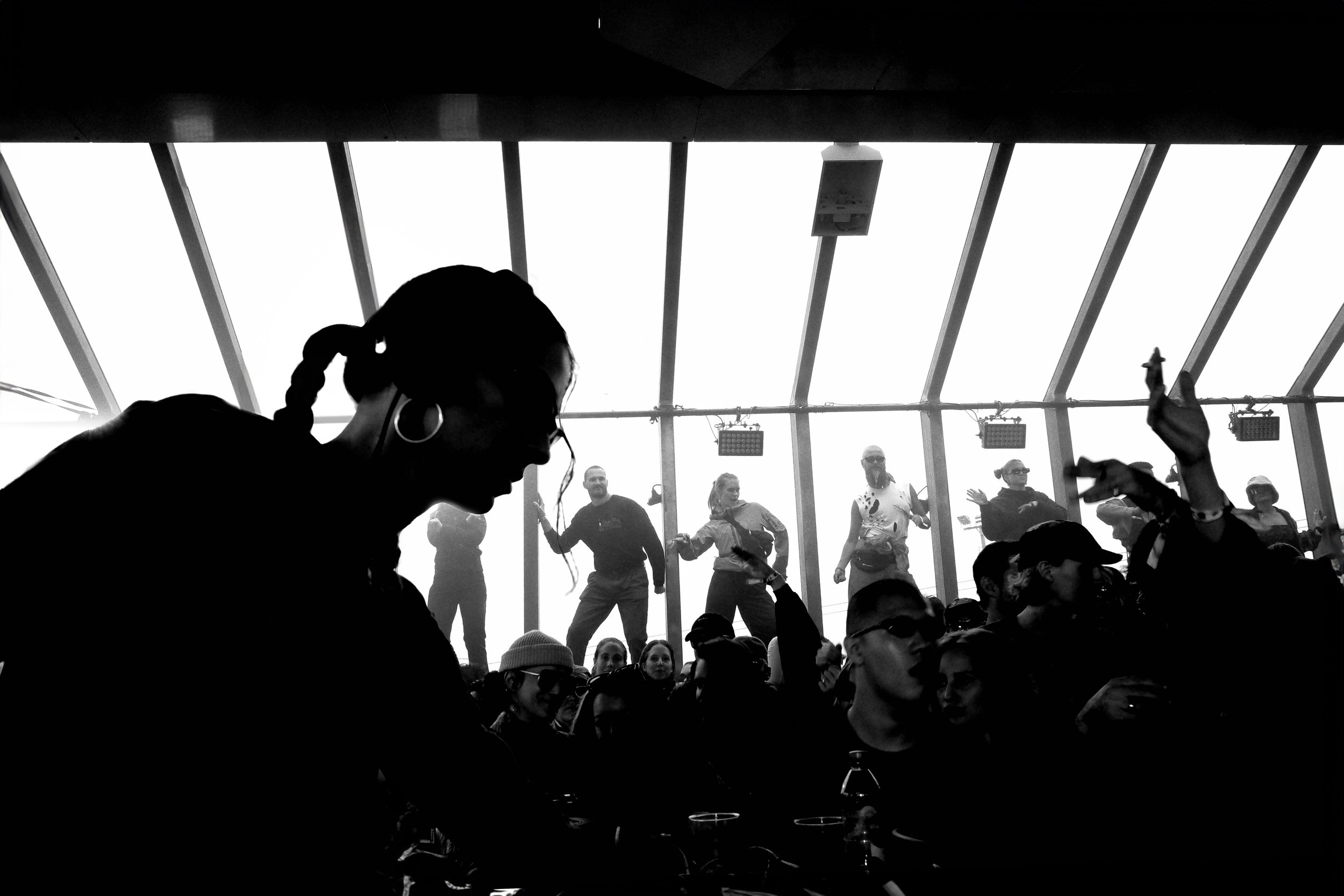 A black and white photo of a crowd inside a large structure with glass walls and ceiling, possibly a greenhouse or atrium, with silhouettes of people dancing and enjoying themselves.