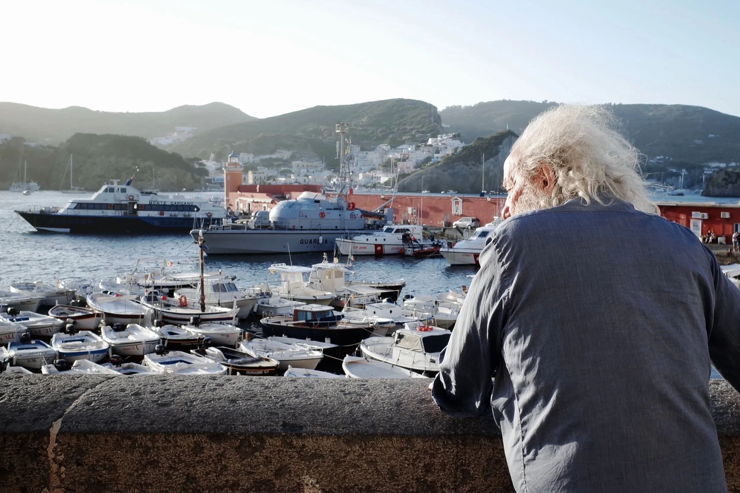 An elderly man with long gray hair sitting by a harbor filled with boats, with hills and white buildings in the background during daylight.