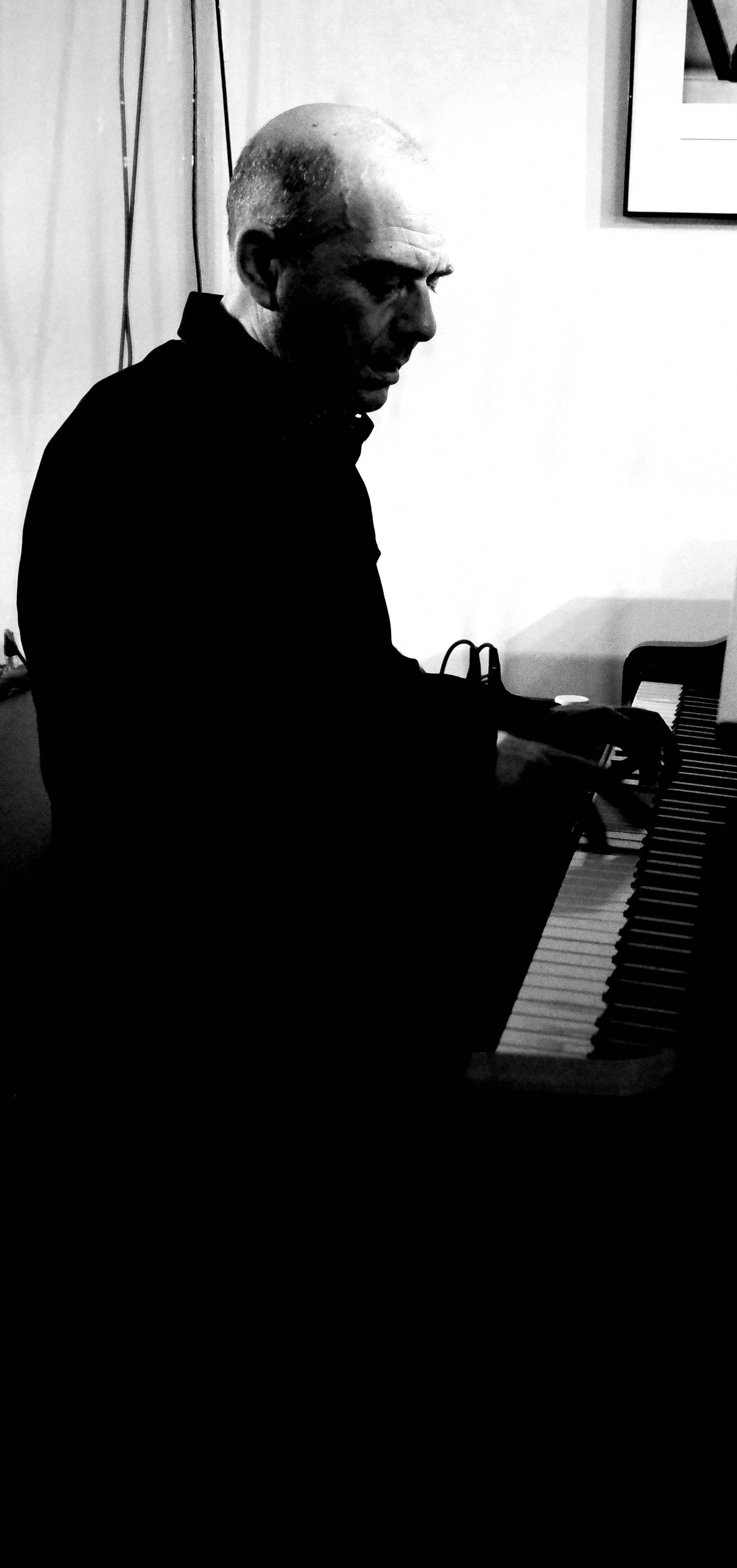 Black-and-white photo of a person playing the piano, with a bald head and dark clothing, focusing on the side profile.
