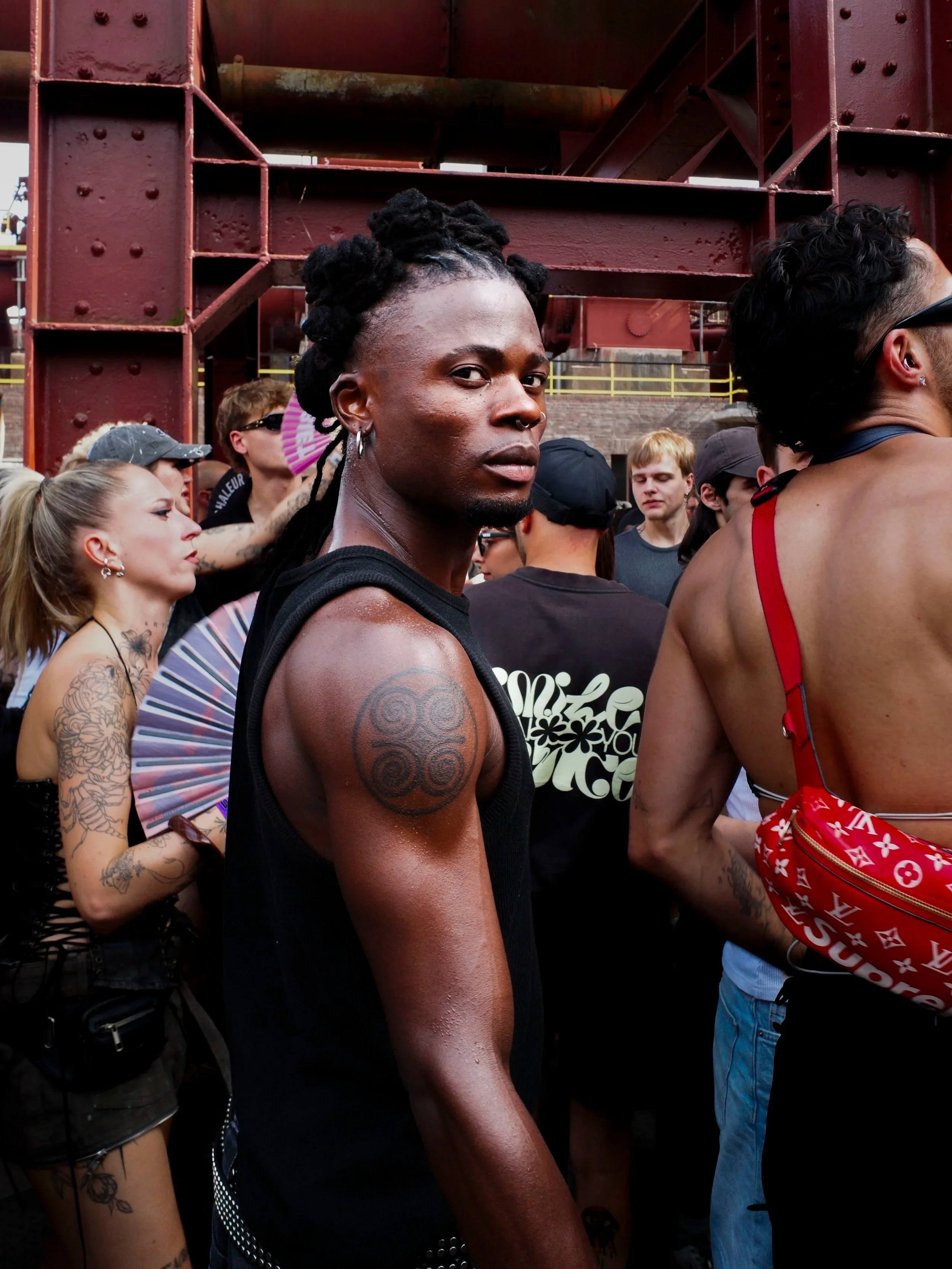 Close-up of a person with dark skin, dreadlocks styled in an updo, tattoos on the arm, and wearing a black tank top, standing amidst a crowd at an outdoor event under a large red metal structure at Stone Techno Festival.