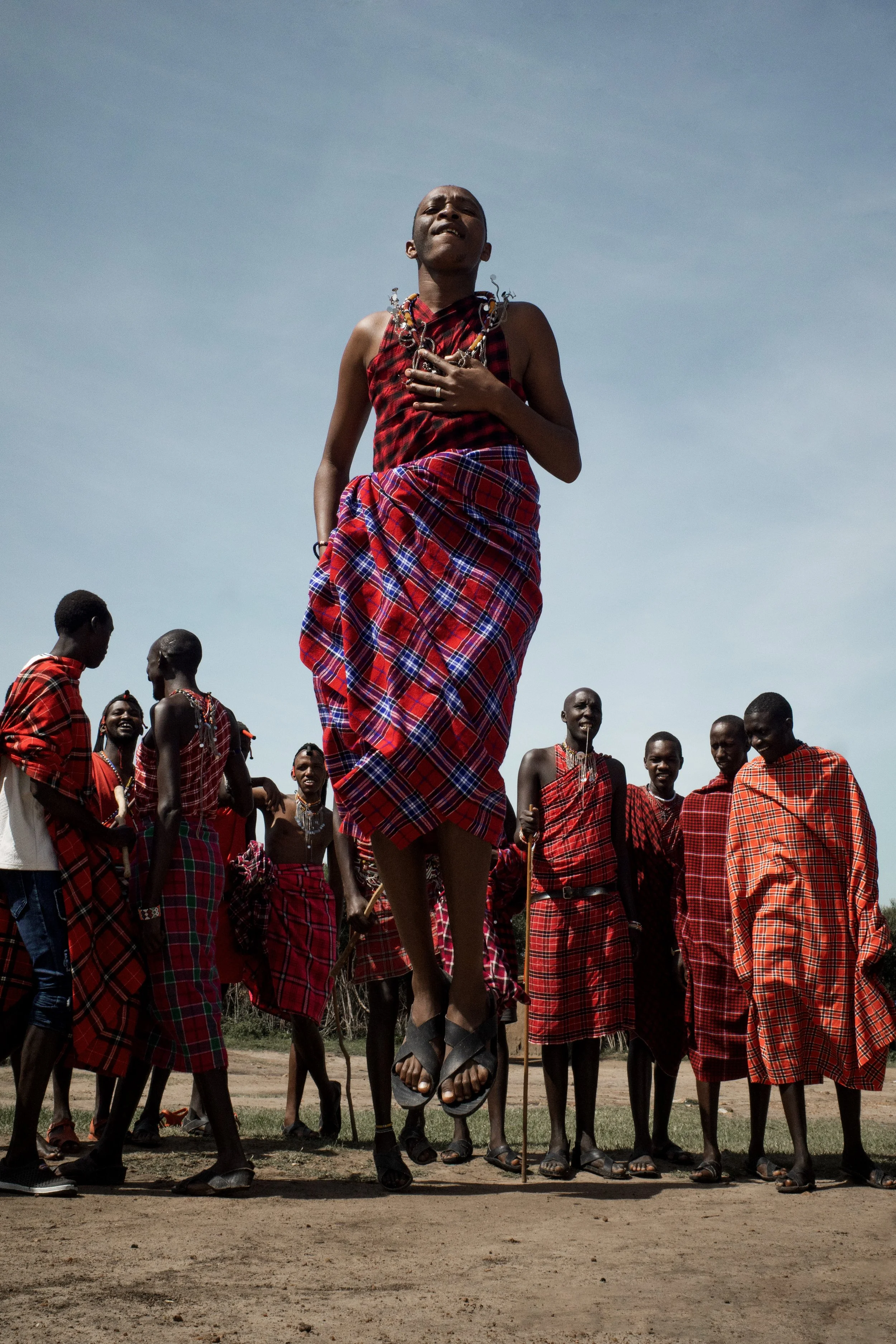 A young Maasai man jumping on a muddy ground, wearing traditional shuka and jewelry, with a group of Maasai people in similar attire standing around him under a clear blue sky.