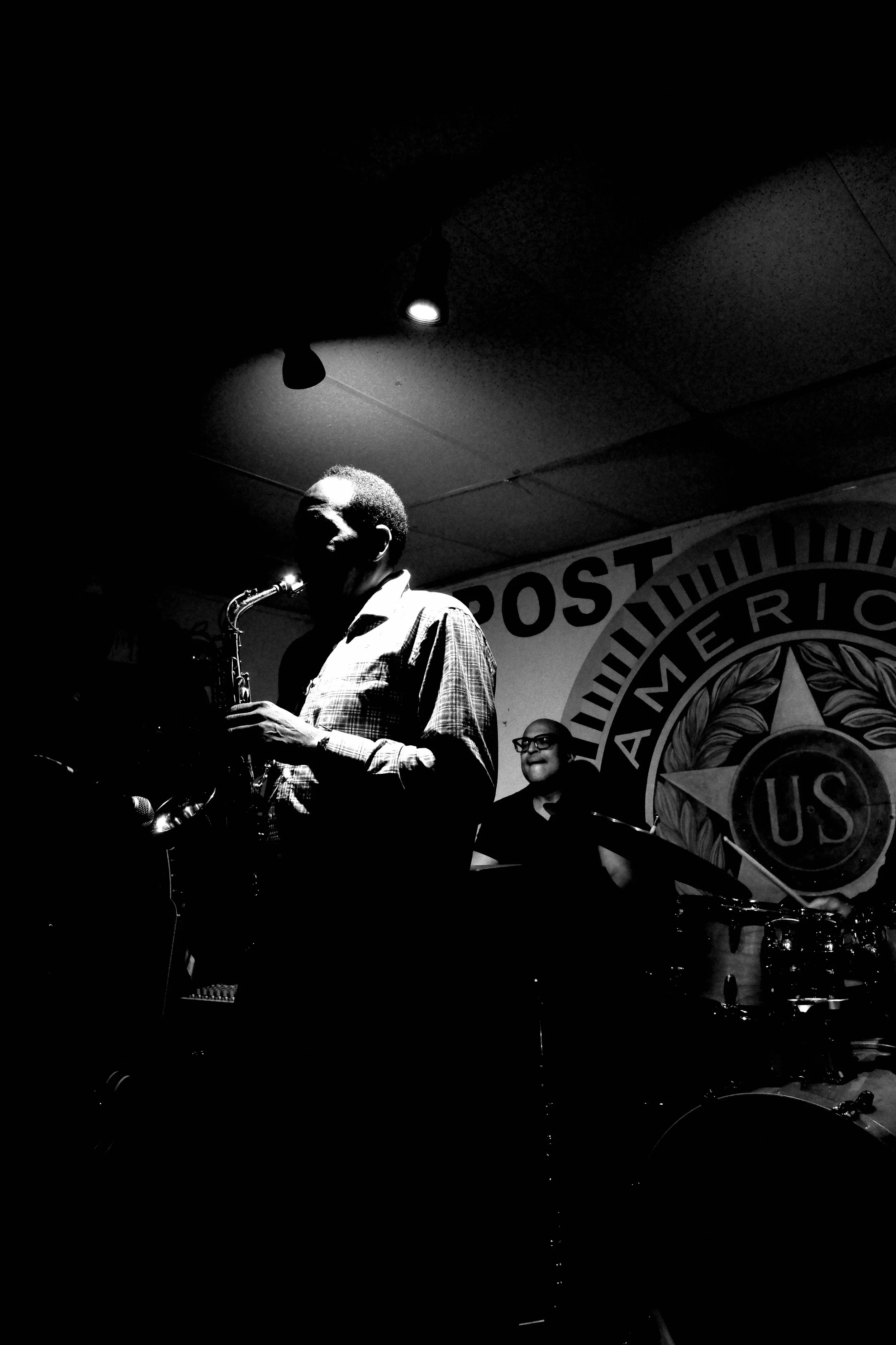 A person playing the saxophone on stage in front of a banner with the U.S. Post Americ logo, with a woman smiling and another person in the background.