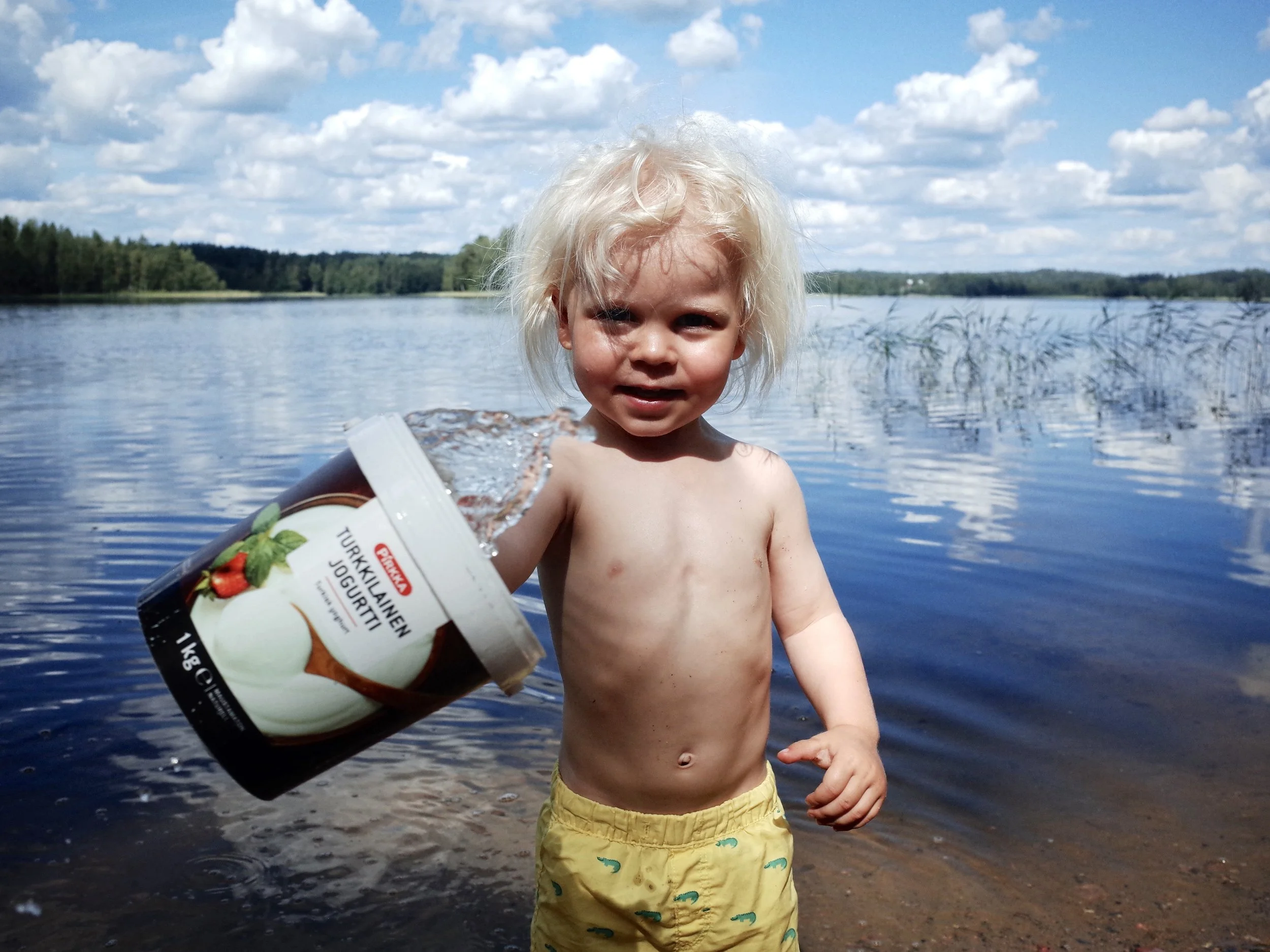 A young child with blonde hair, wearing yellow shorts with a dinosaur pattern, playing in a body of water on a sunny day with a partly cloudy sky. The child is holding a container of yogurt and smiling at the camera, surrounded by a natural landscape with trees in the background.