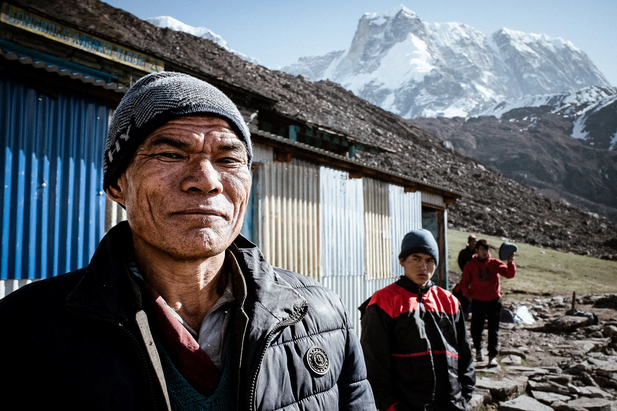 A man in a beanie and jacket standing outdoors in a mountainous area with snow-capped peaks in the background. Other people are visible behind him, dressed in outdoor gear.