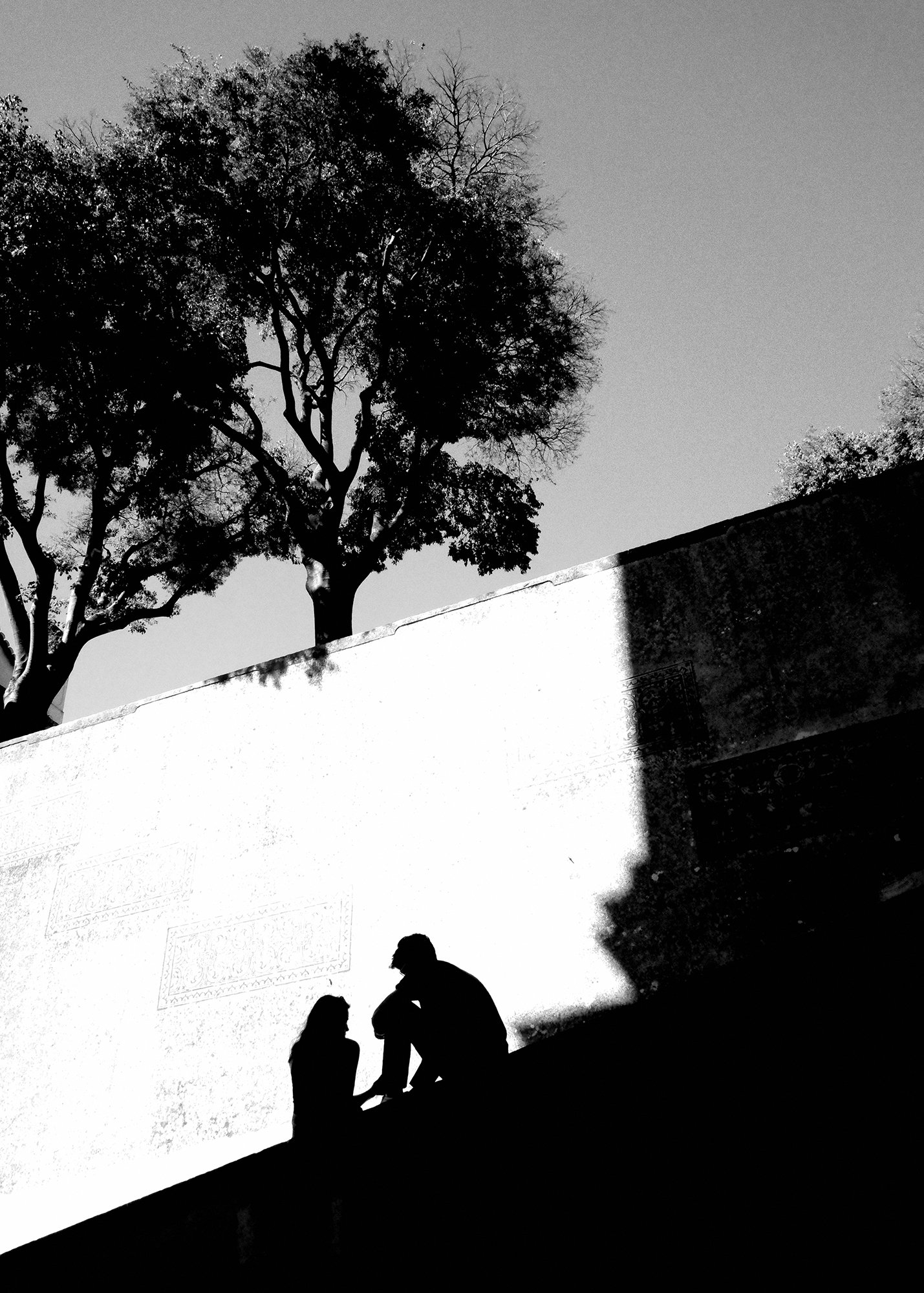 Silhouettes of three people outdoors against a wall with trees in the background, one person sitting and two standing close together, engaging in conversation.