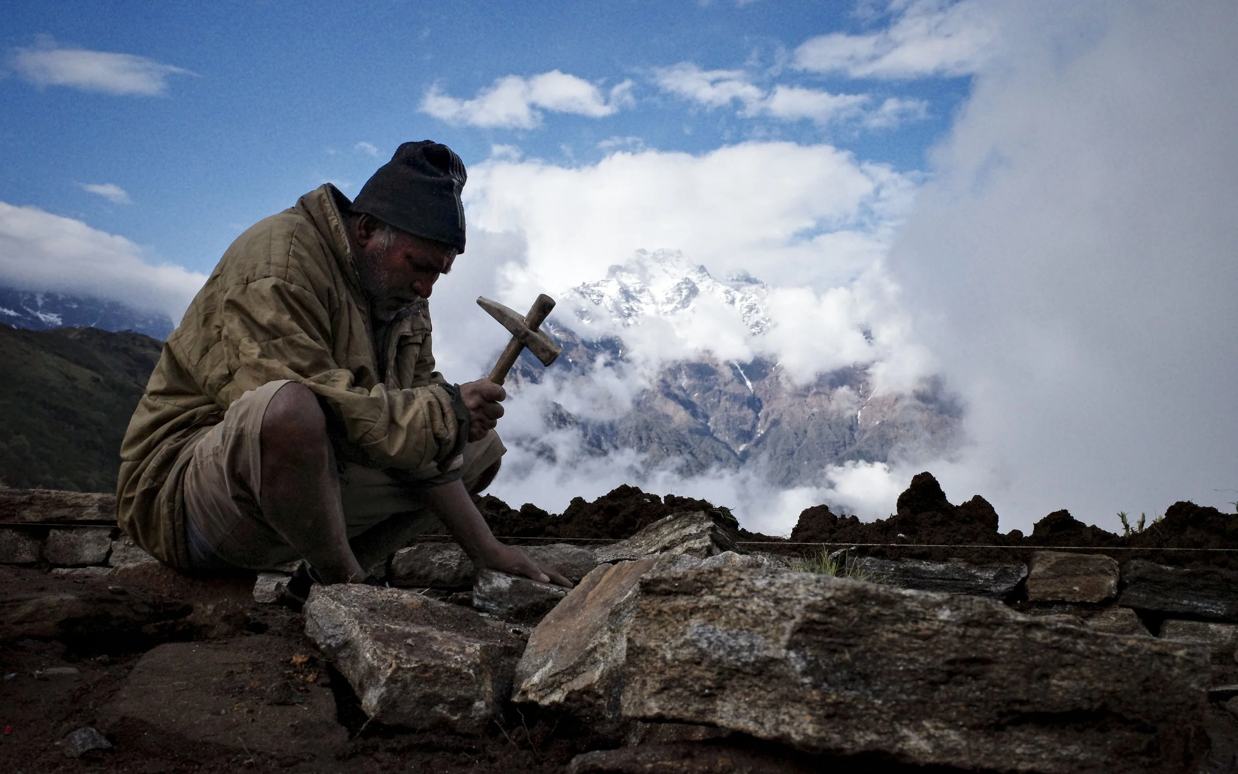 A person dressed in warm outdoor clothing, holding a hammer and working on a rocky surface with a mountain landscape and partly cloudy sky in the background.