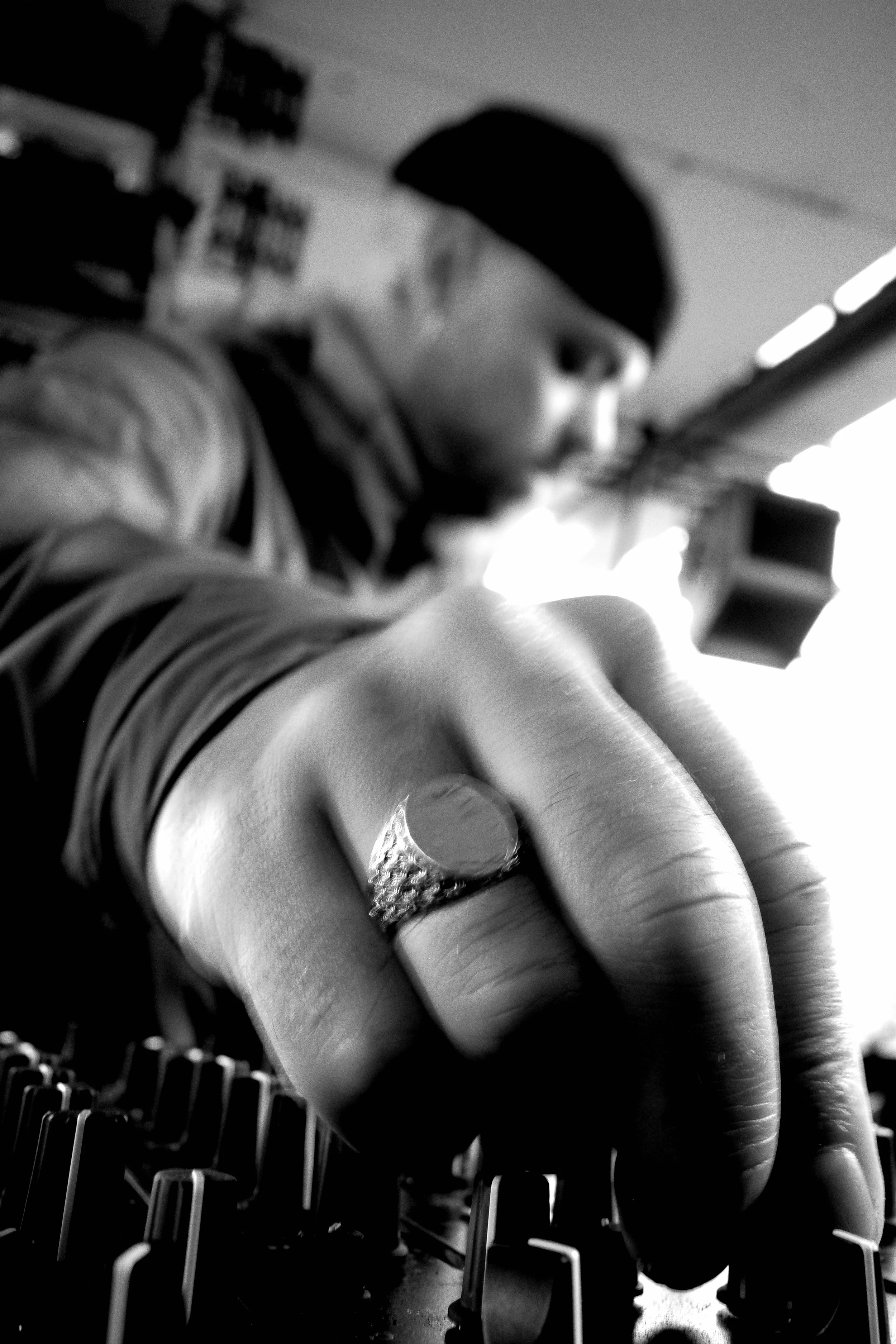 Close-up black and white photograph of a person’s hand wearing rings, reaching out towards the camera with chairs in the background.