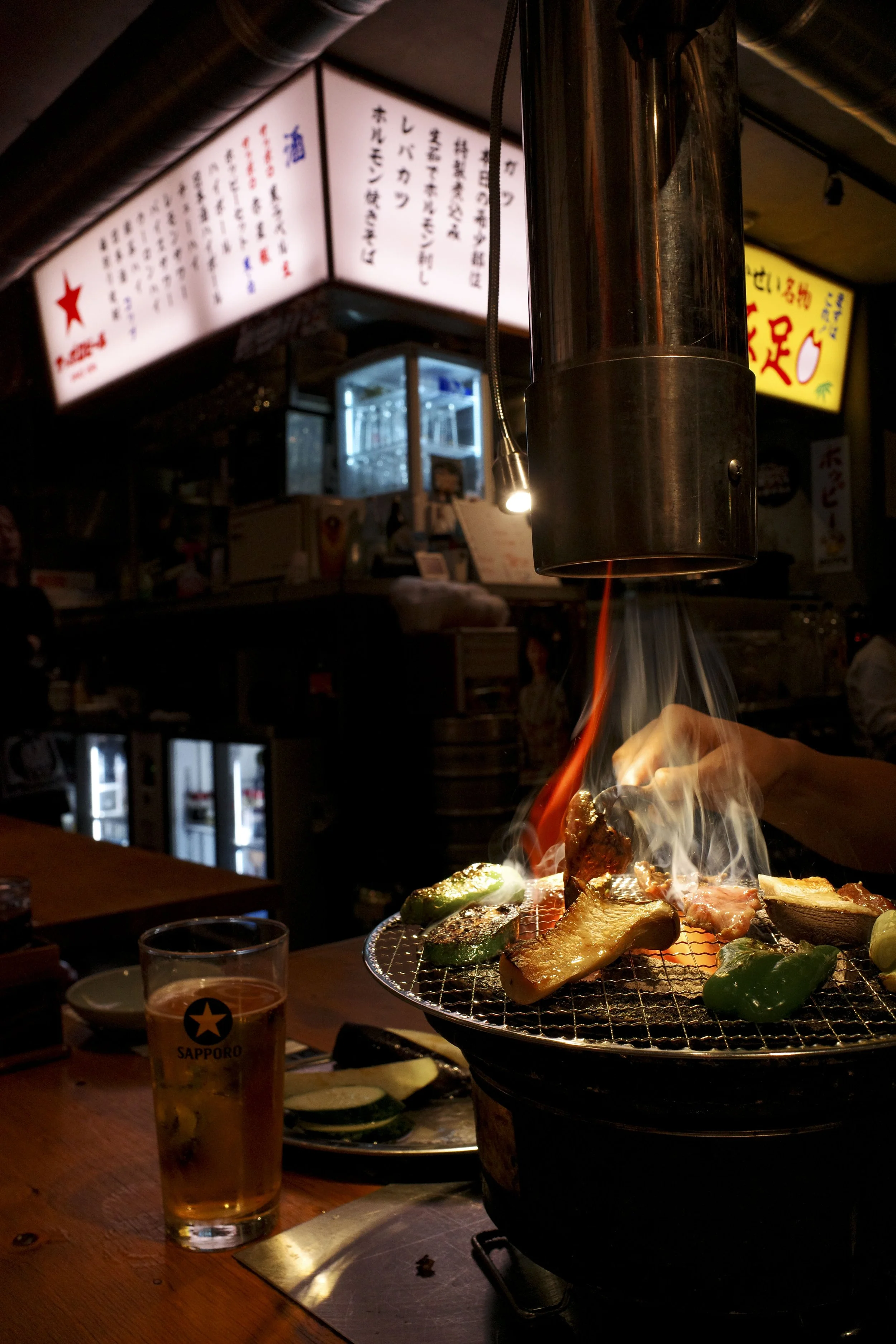A grill with various meats and vegetables cooking, with smoke and flames, inside a restaurant. A glass of Sapporo beer is on the table near the grill. Menus with Japanese writing are visible in the background in Tokyo, Japan.
