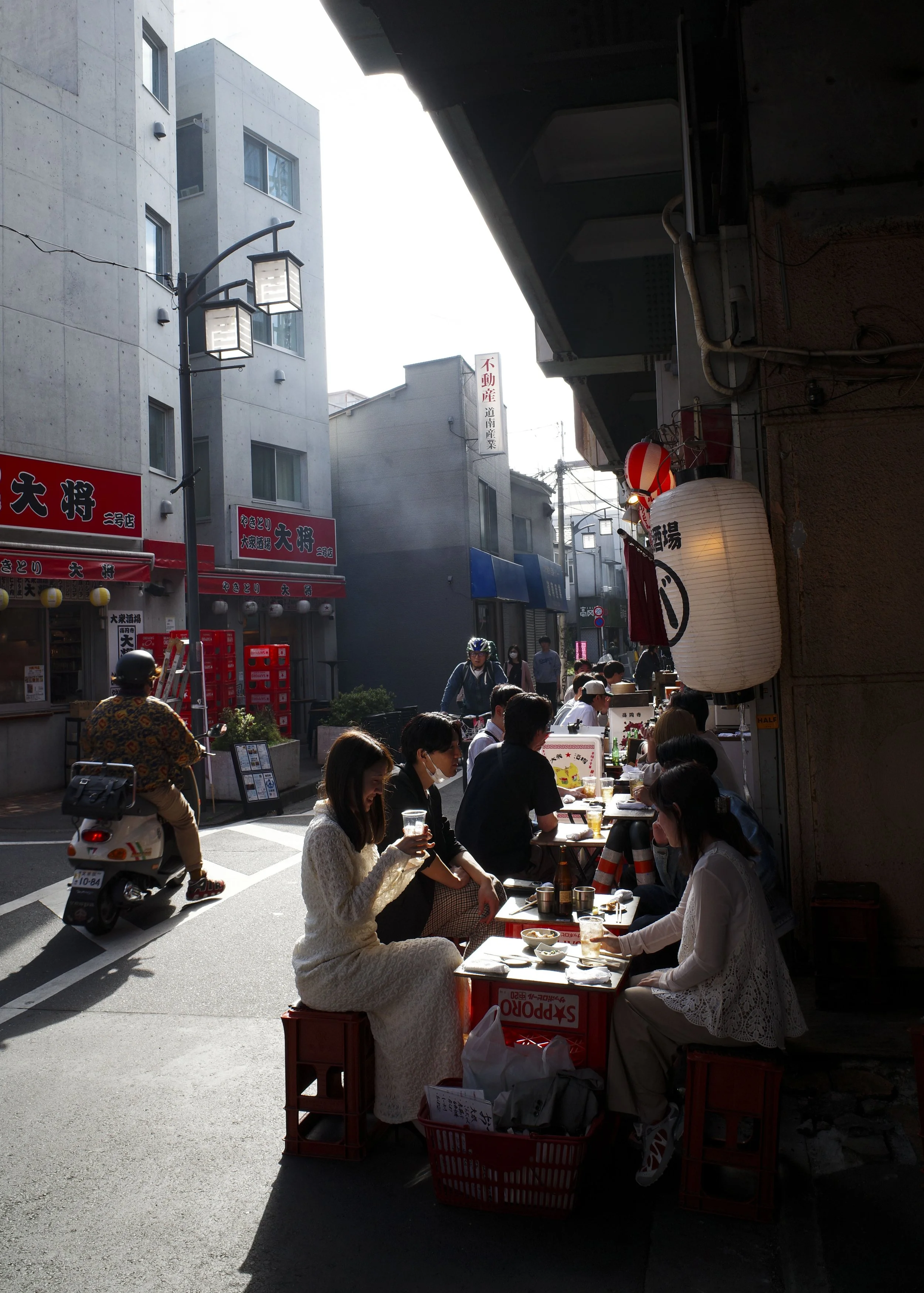 People sitting at outdoor tables on a city street, with traditional Japanese paper lanterns hanging above, in Tokyo, Japan.