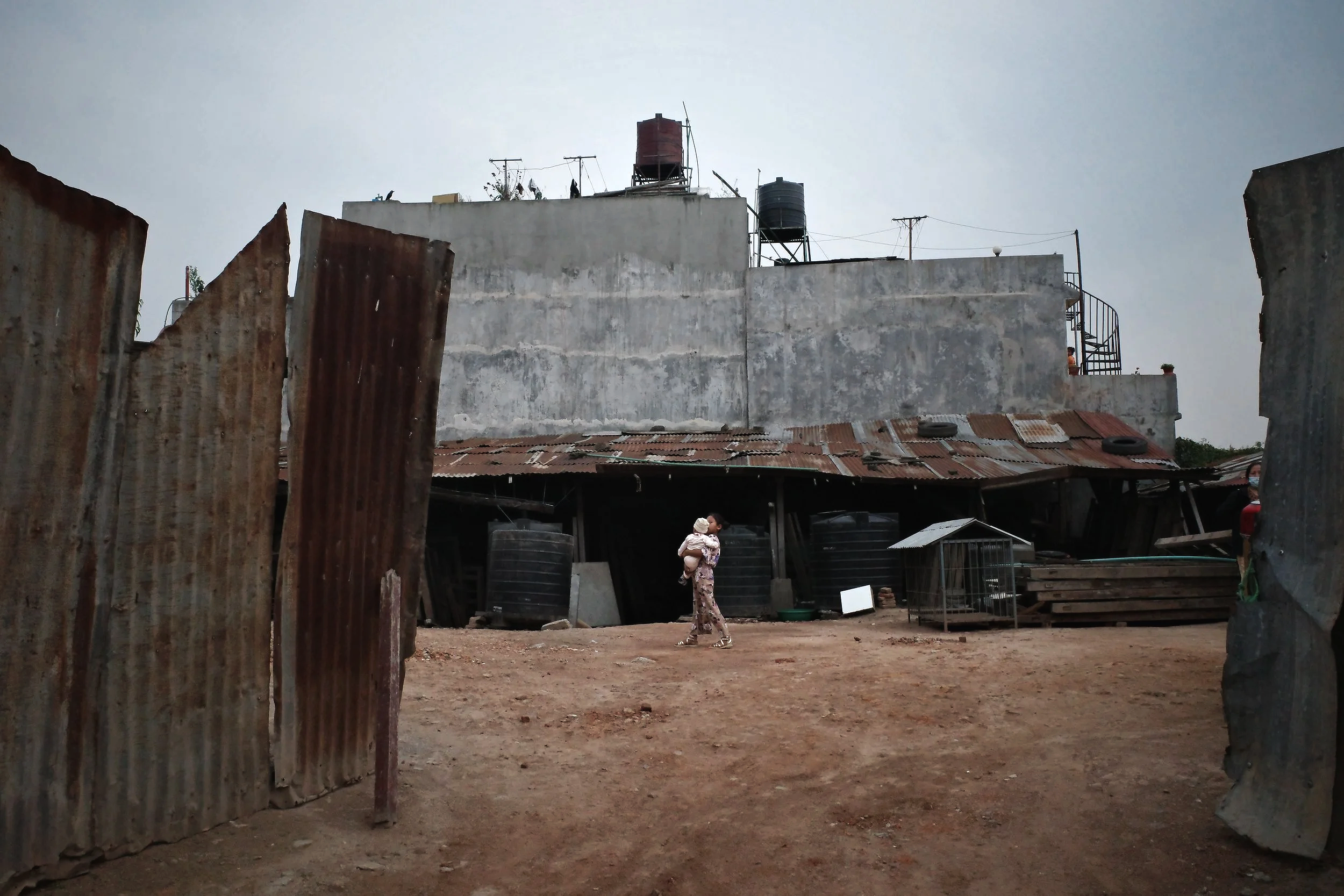 A woman holding a child walking in a dirt yard, with a backdrop of concrete and metal buildings and corrugated metal fences, in a developing area.