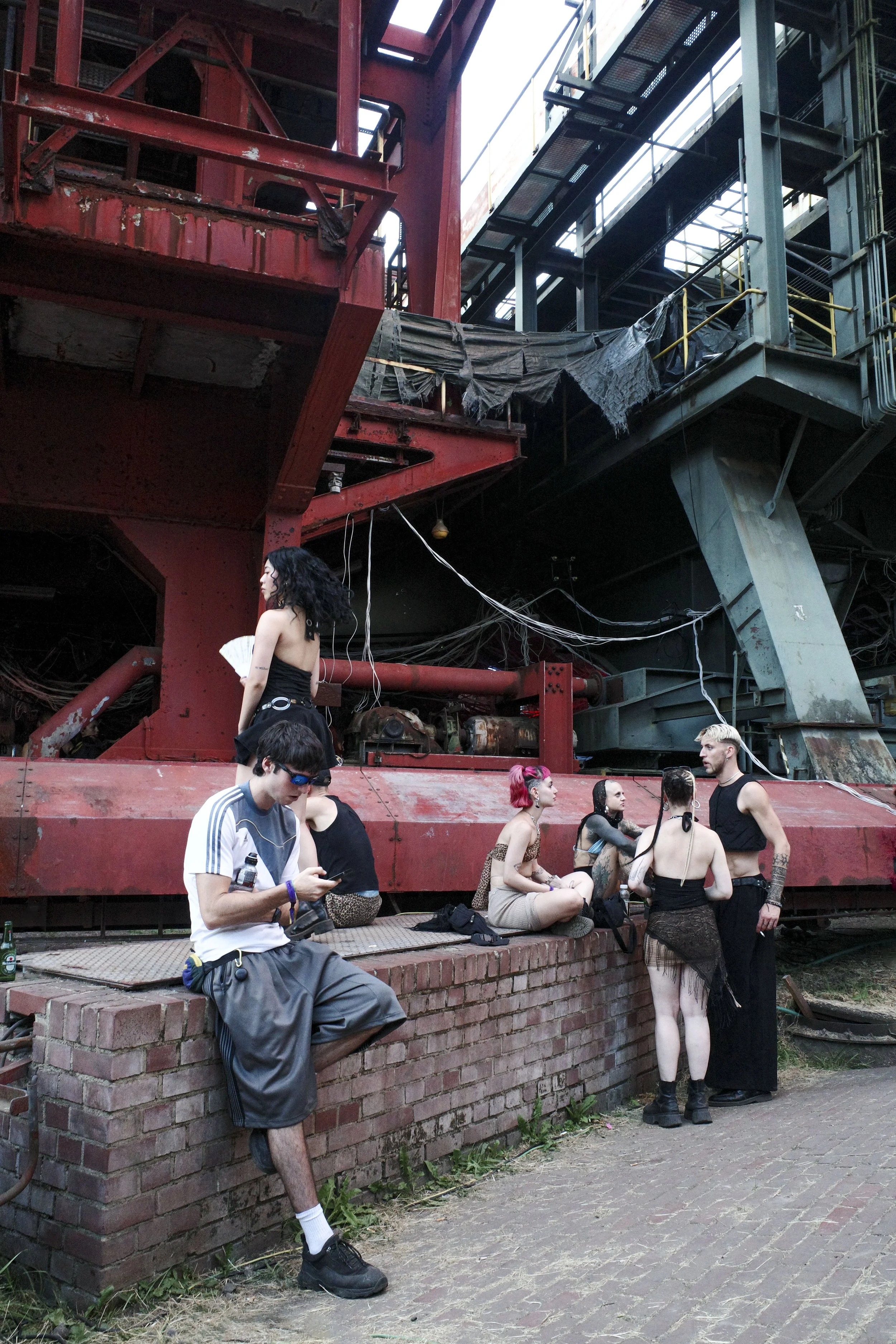 People gathered outdoors near an industrial steel structure, some sitting on a brick ledge and others standing, with diverse clothing styles and hairstyles at Stone Techno Festival..