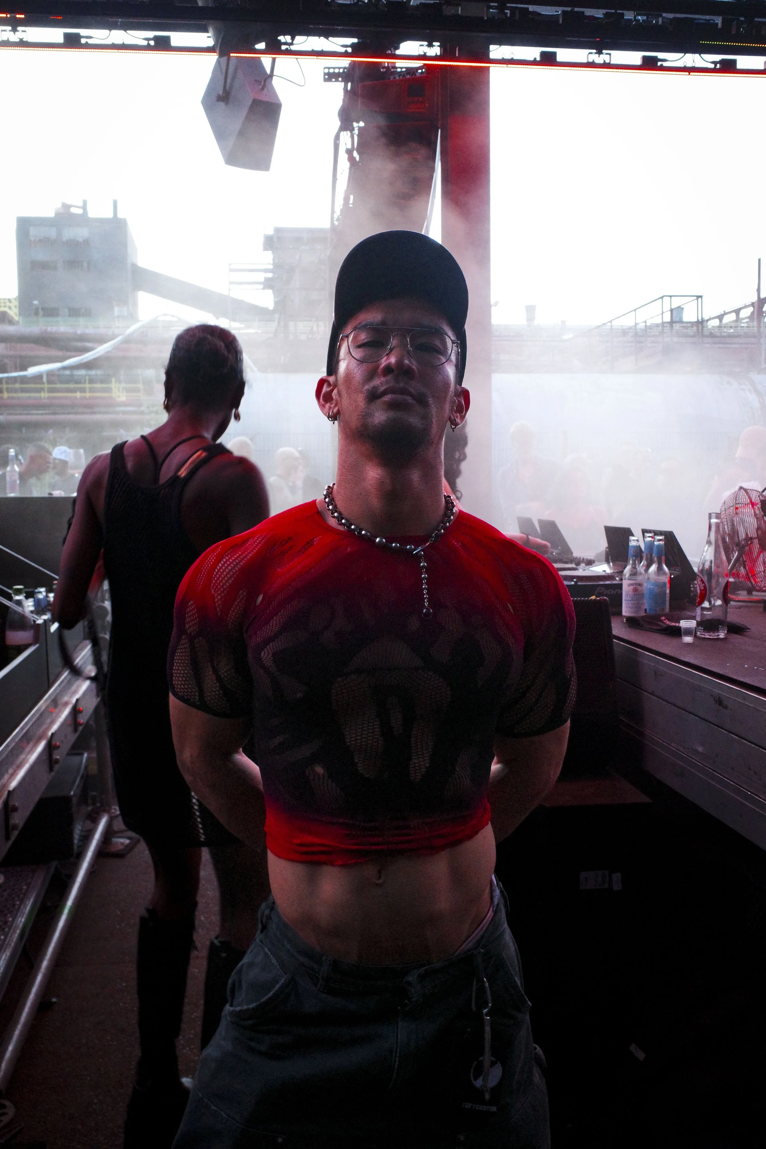 A man with glasses, earrings, and a chain necklace, wearing a black cap, red mesh shirt, and dark jeans, standing with arms behind his back at a music event, with DJ equipment and other people in the background at Stone Techno Festival.