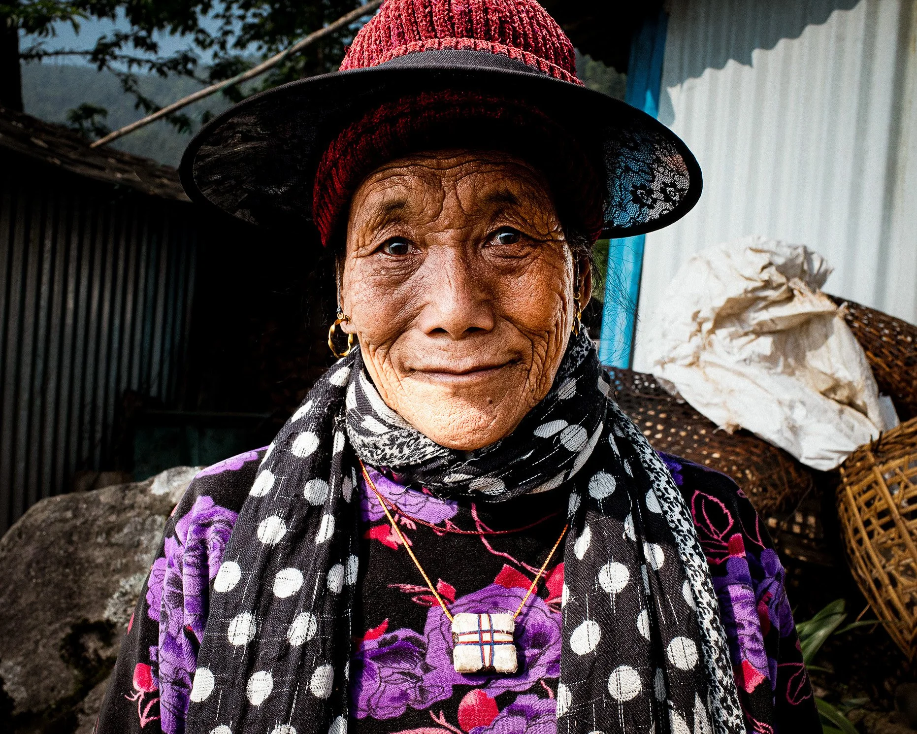 Close-up of an elderly woman with wrinkled skin, wearing a red knit hat, black lace hat, gold earrings, a necklace with a cross pendant, a purple floral top, and a black scarf with white polka dots. She is outdoors with a house or shed and a pile of white sacks visible in the background.