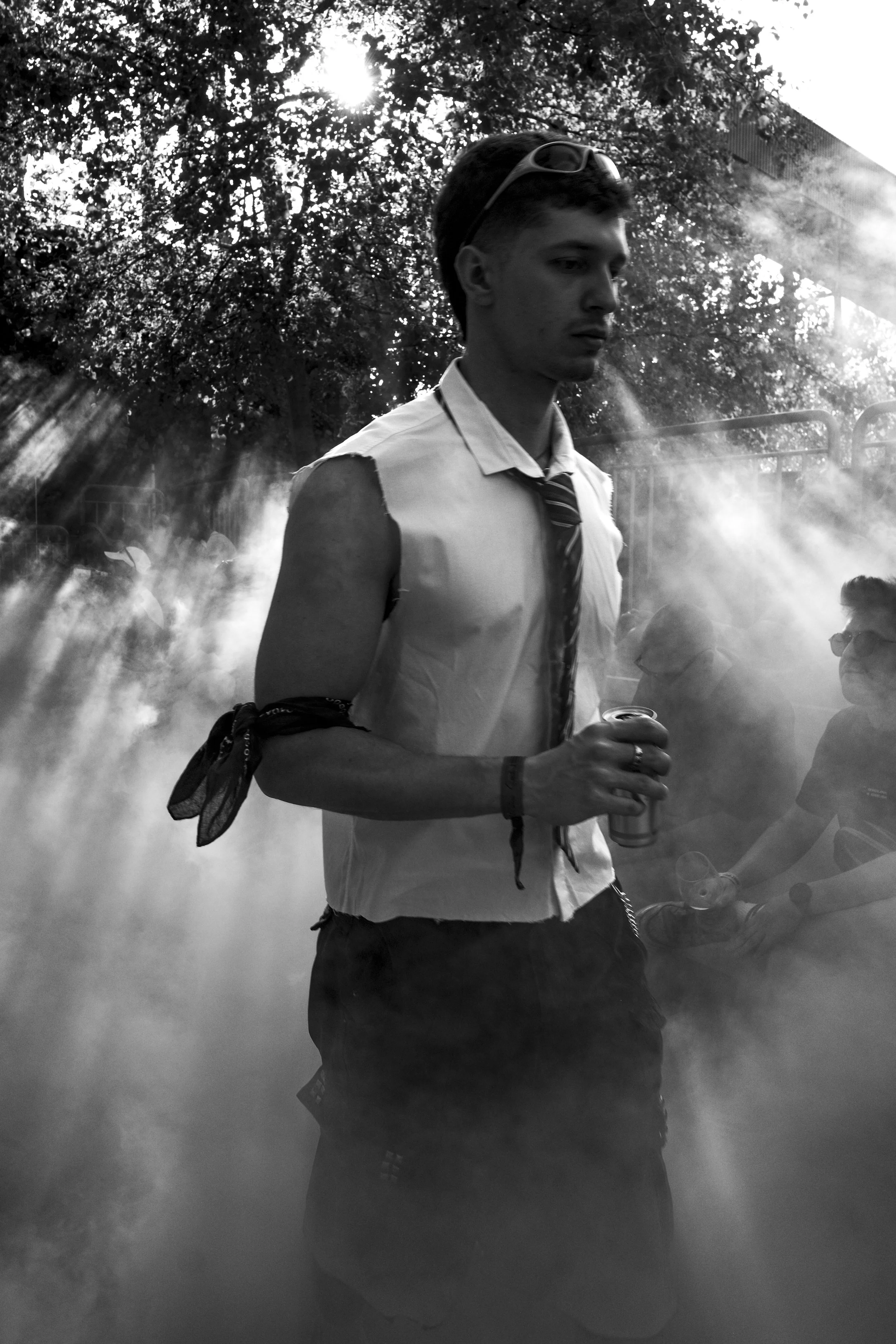 A young man wearing sunglasses, a sleeveless shirt, and a tie, holding a drink, standing outdoors in front of a background with trees and sunlight, surrounded by mist or smoke at Stone Techno Festival.