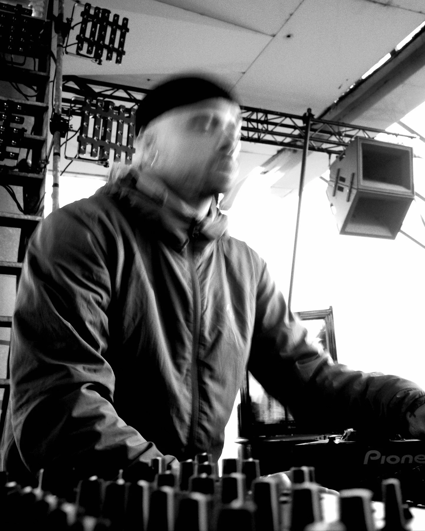 A person wearing a jacket and headphones, possibly a DJ, standing behind DJ equipment in a room with lighting and stage equipment.