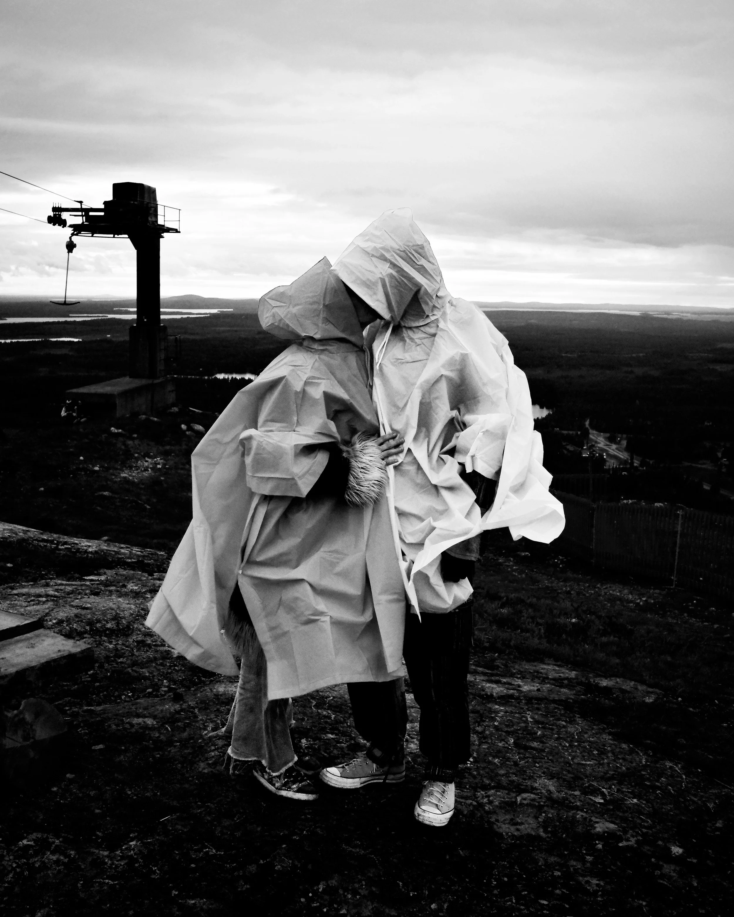 Two people in white protective suits embracing each other outdoors under a cloudy sky, with a landscape of fields and water in the background and a crane nearby.