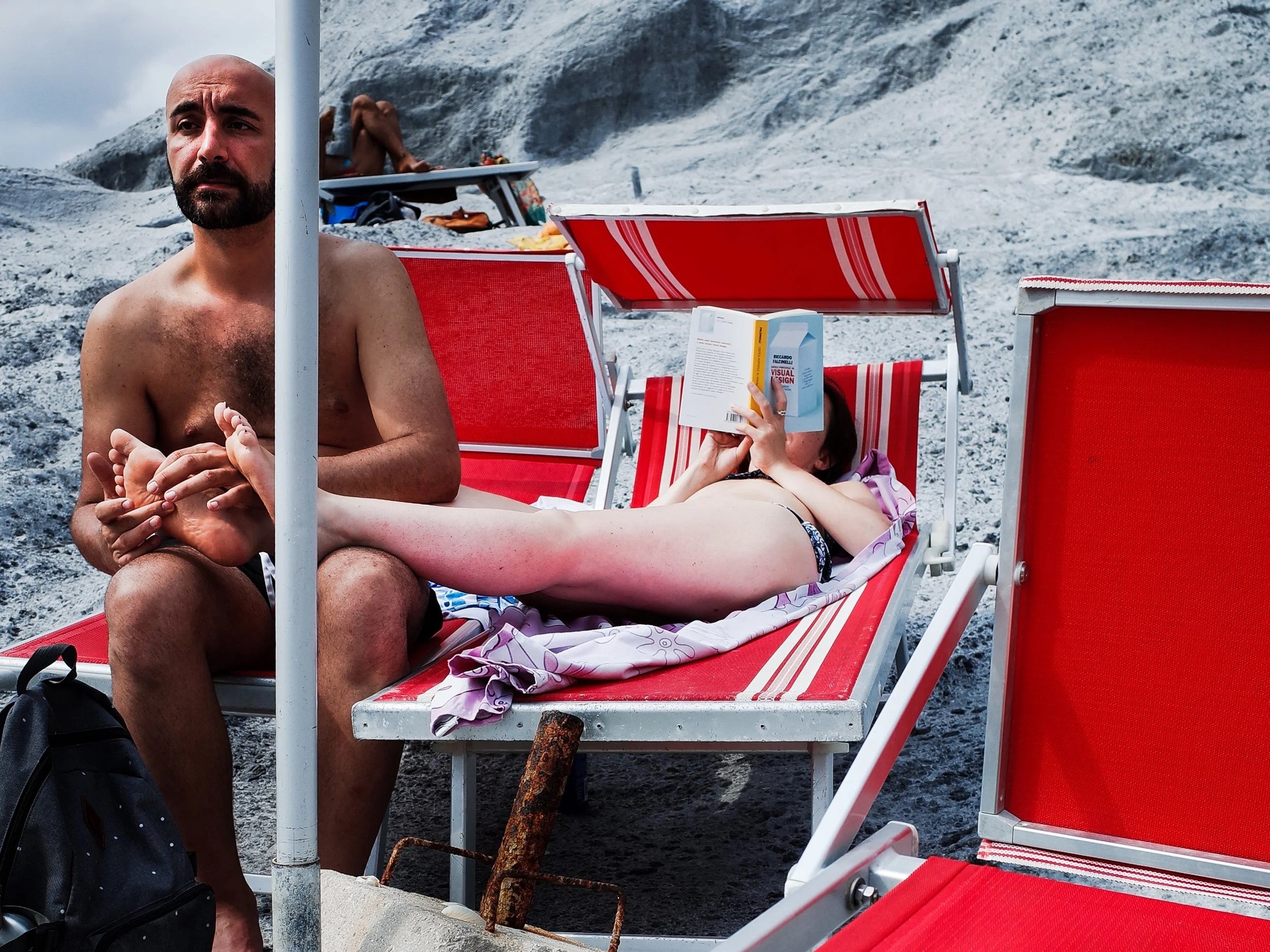A shirtless man with a beard and a bald head sitting on a red beach chair at the beach, holding a woman’s foot who is lying on the chair and reading a book on a rocky beach in Ponza, Italy. In the background, there is another person sitting.