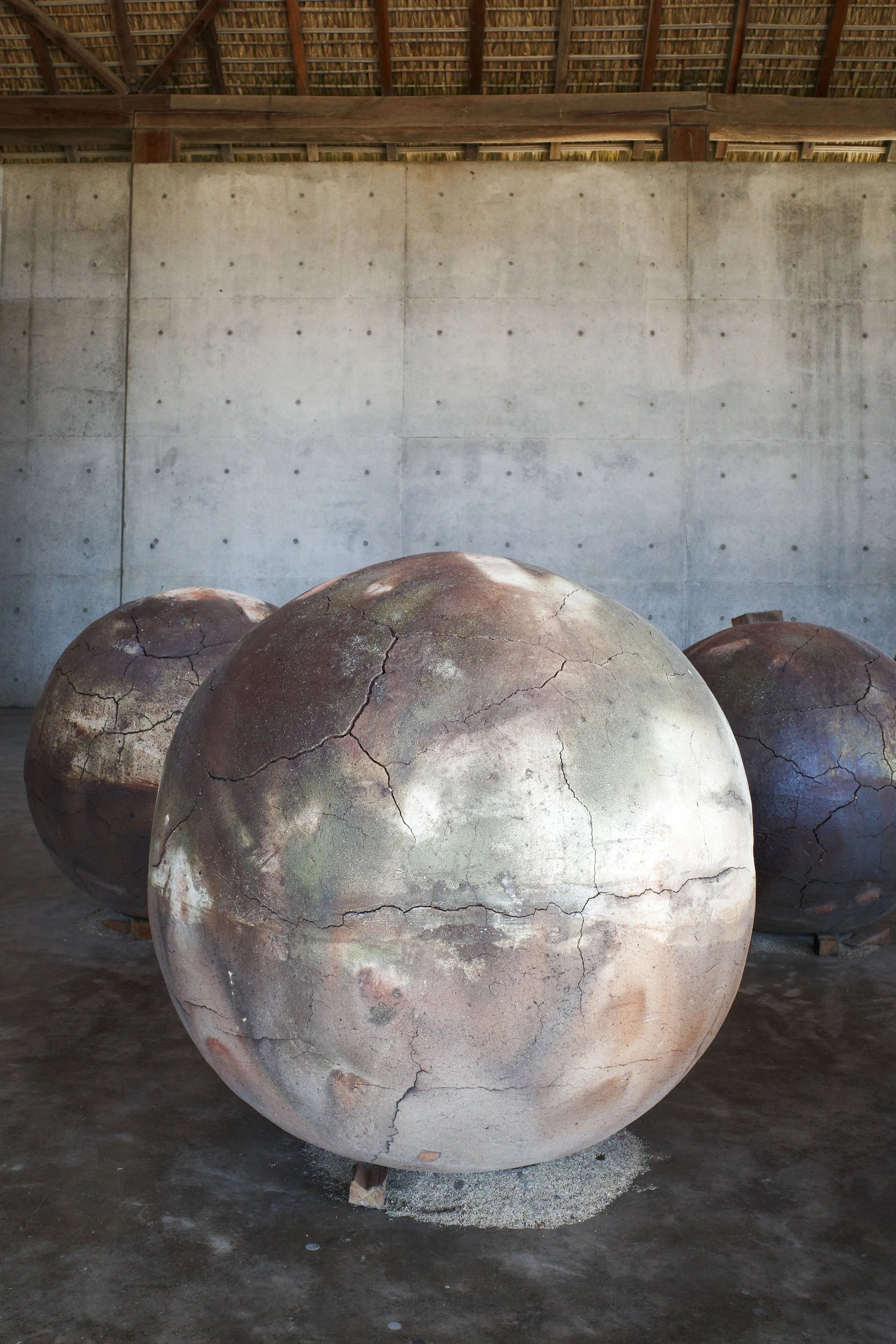 Large clay pots with cracks, placed on a concrete floor inside a structure with a thatched roof and a concrete wall in Casa Wabi in Puerto Escondido.