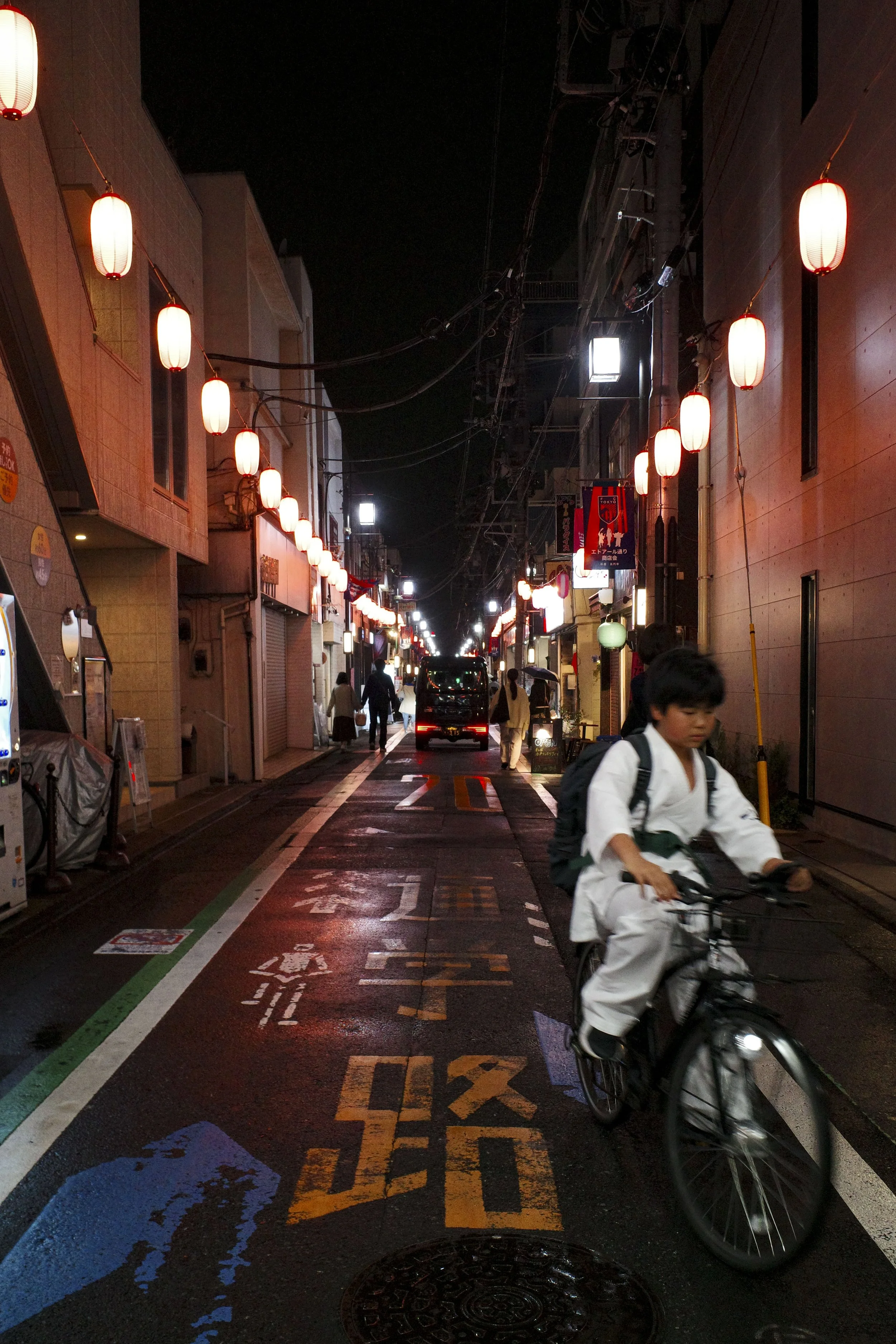 Nighttime street scene in Japan with red paper lanterns hanging above, people walking, and a person riding a bicycle in a white traditional outfit in Tokyo, Japan..