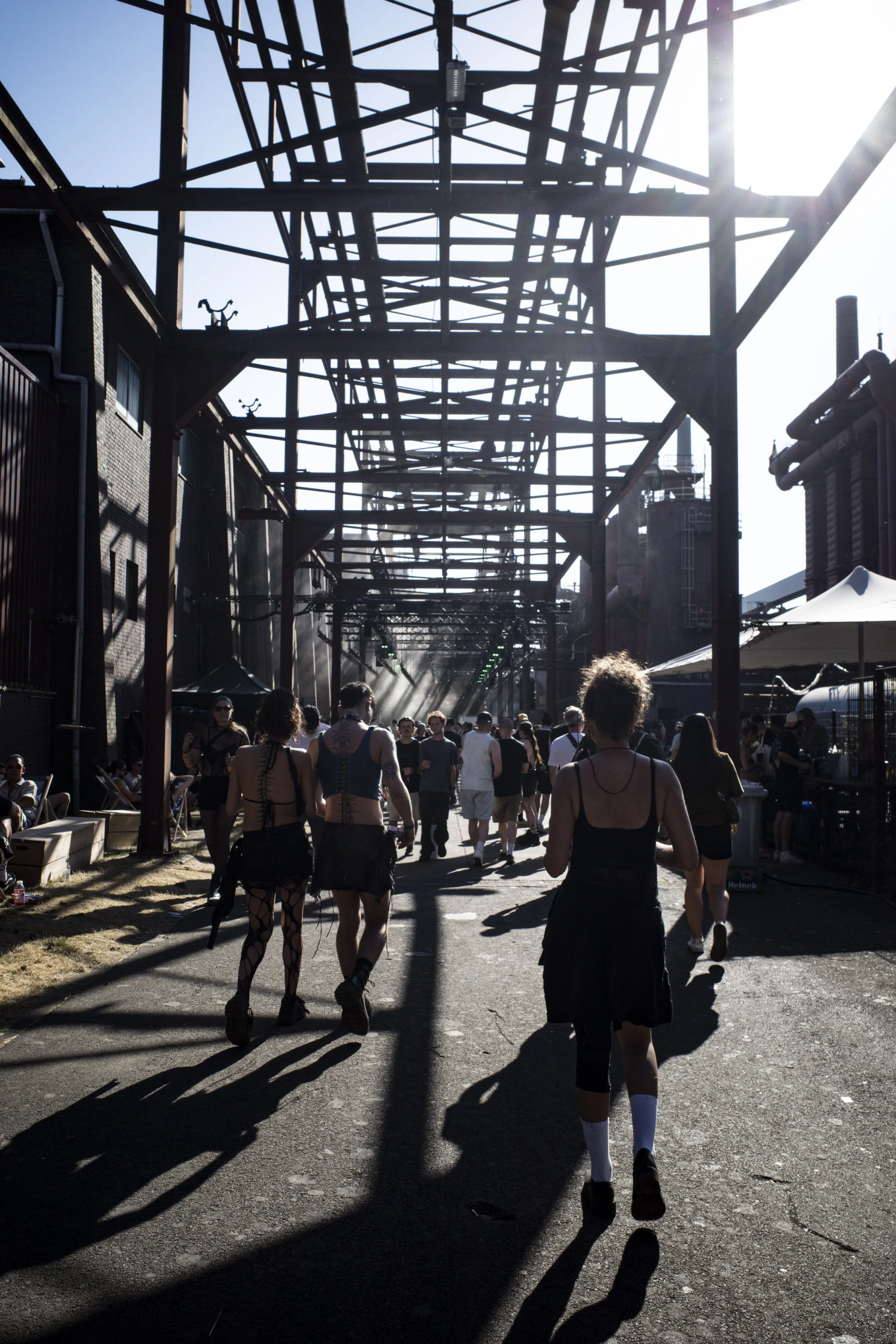 People walking outdoors under metal structures with industrial chimneys and tents in the background, sunny weather casting long shadows at Stone Techno Festival stage.