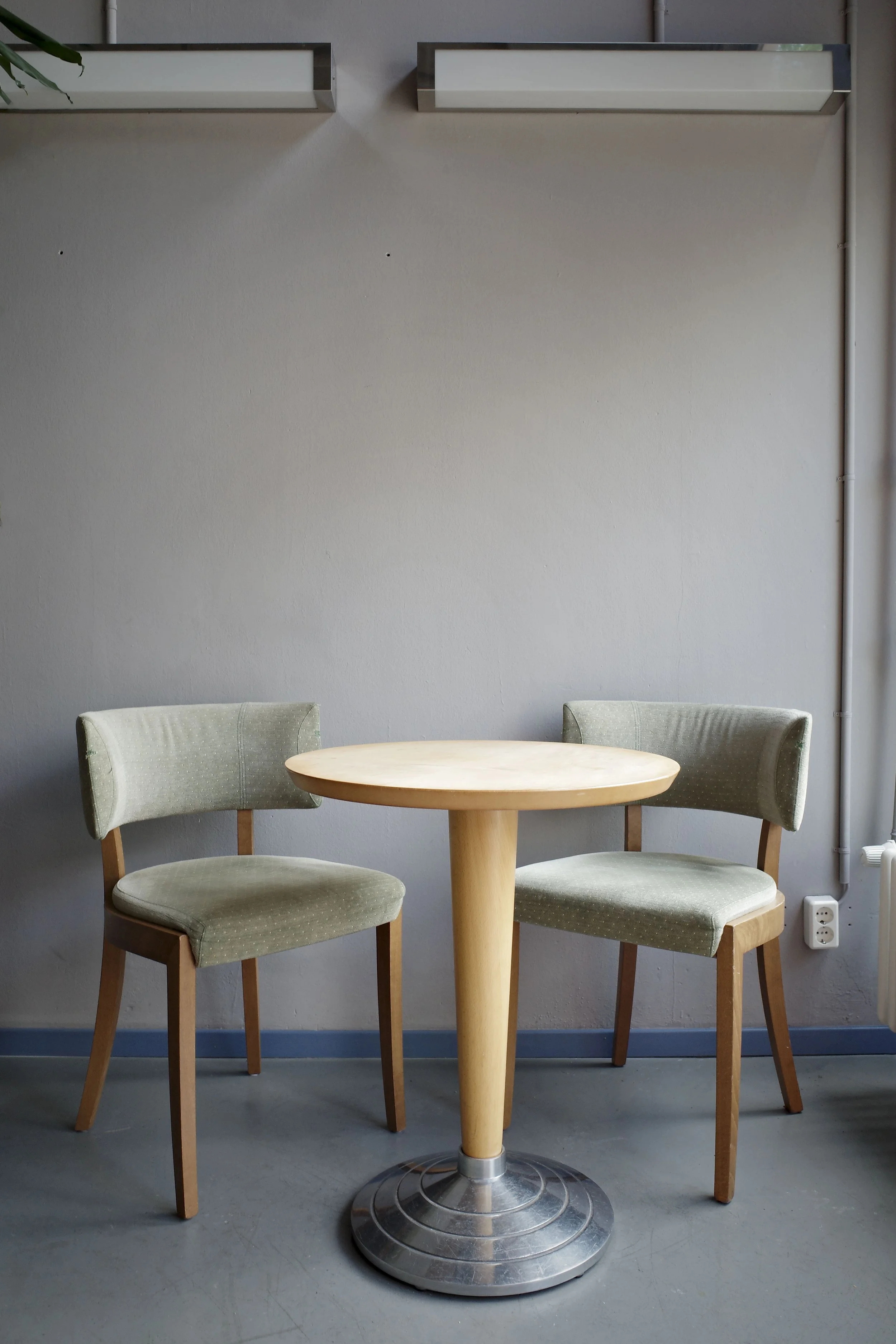A round wooden table with a metal base, surrounded by three green upholstered chairs with wooden legs, set against a gray wall with two horizontal light fixtures and a small section of a window with a plant in the top left corner.