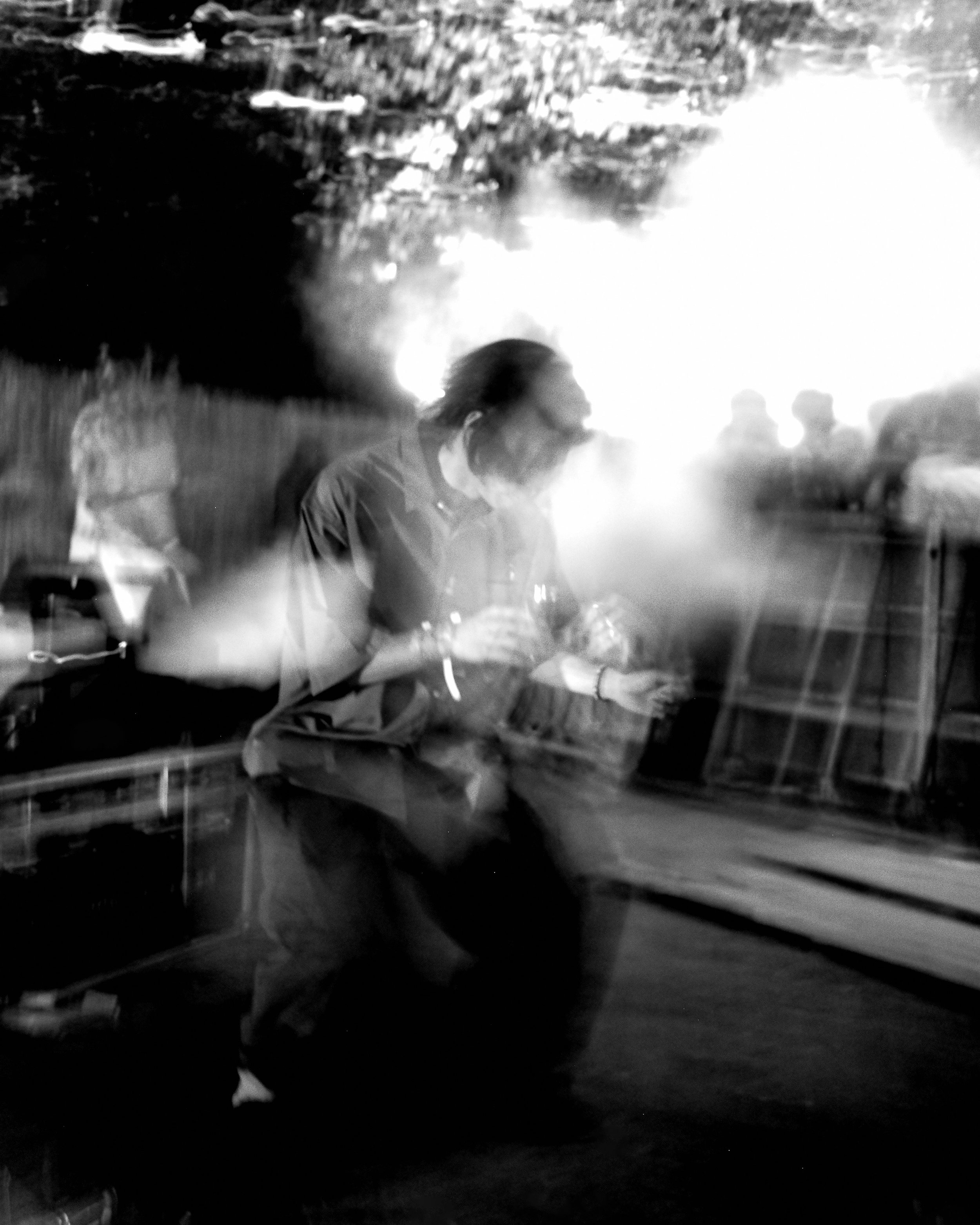 Black and white photo of a person with dark hair and glasses sitting at a table, with a large bright light or reflection in the background, along with cars and trees outside the window.