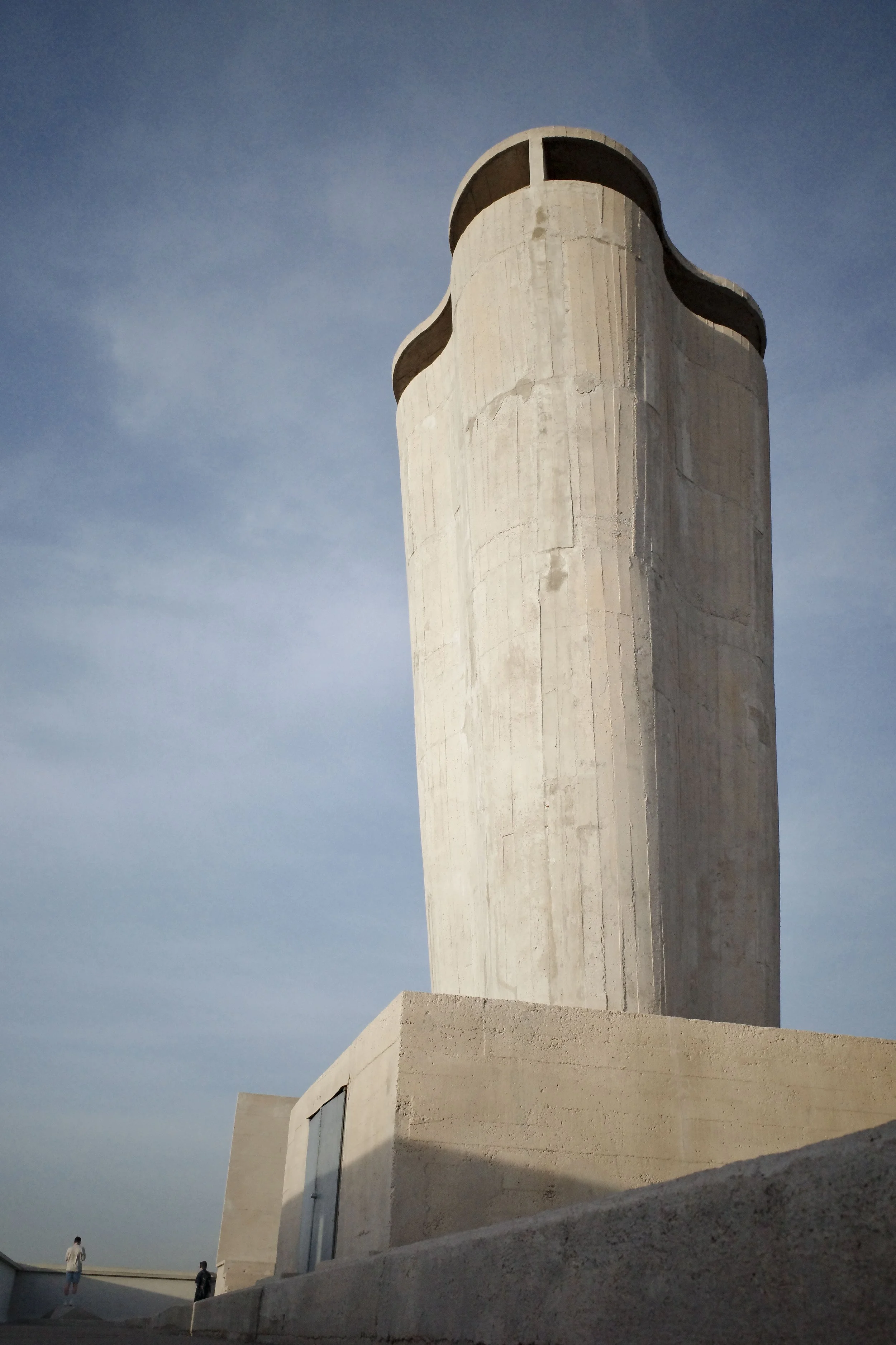 A modern, minimalist white building with curved edges and large glass windows designed by Le Corbusier, set against a clear blue sky, with a flat rooftop and a few people standing on the ground nearby in Marseilles, France.