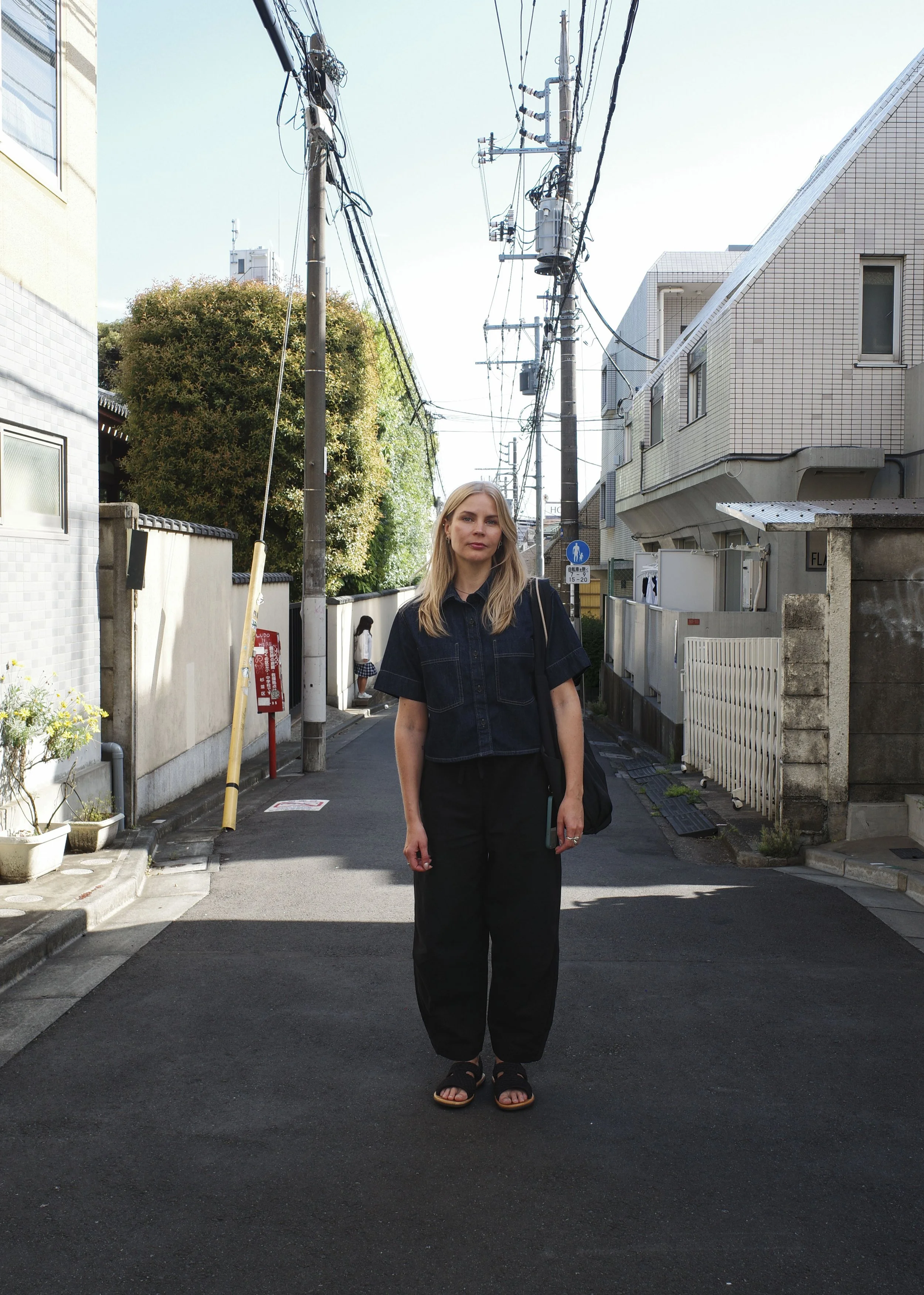 A young woman with blonde hair, wearing a black denim shirt, black pants, and black sandals, stands on a narrow street in front of a fence and residential buildings under a clear sky in Tokyo, Japan.