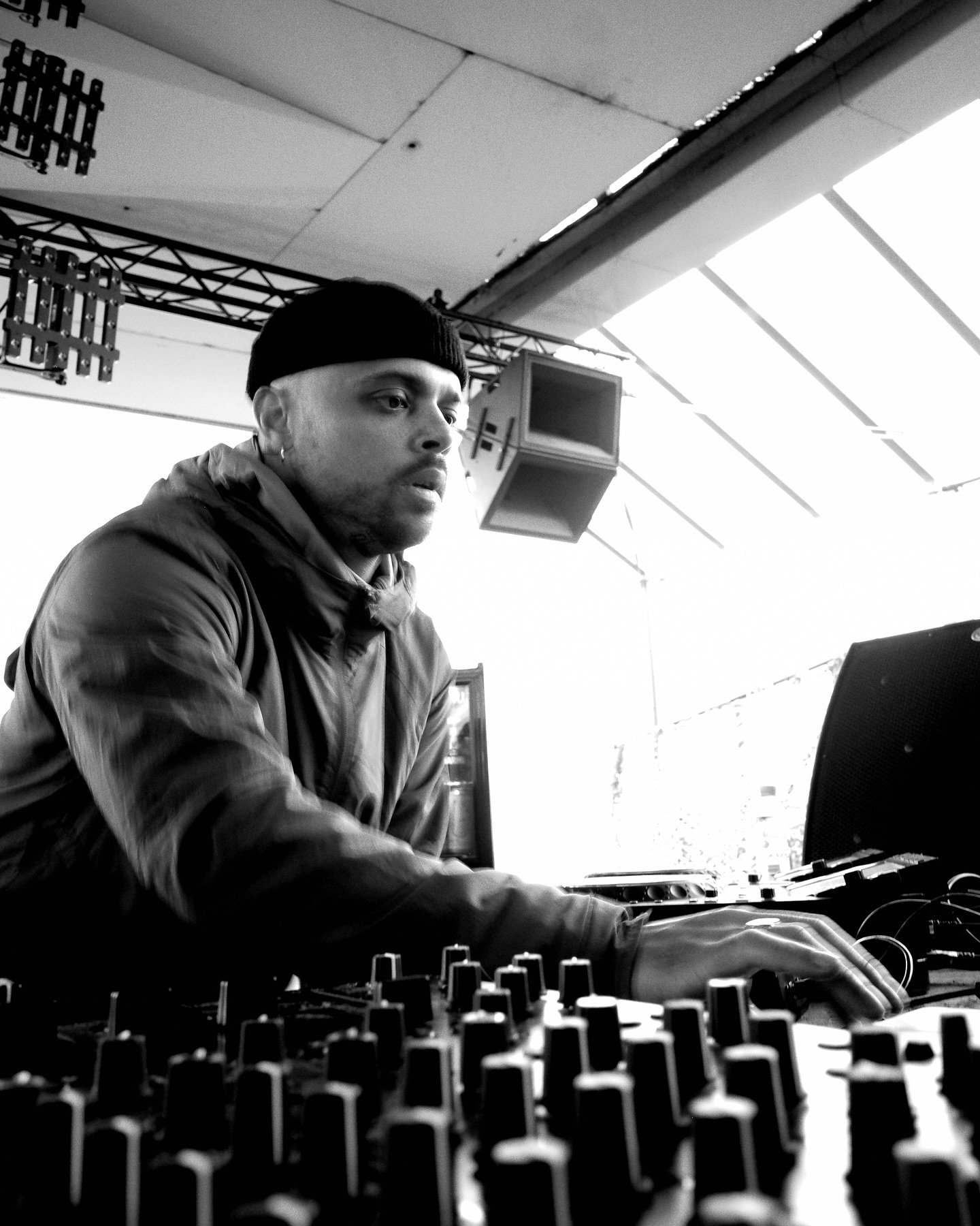 A man wearing a black beanie and jacket operating DJ equipment, with a large outdoor stage and lighting setup in the background.