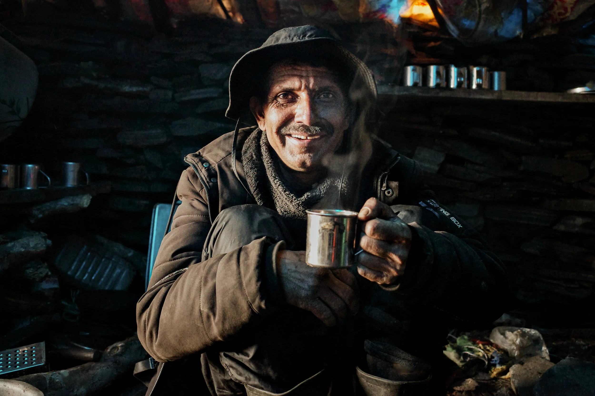 A man with a big smile, wearing a hat and warm jacket, sitting in a dark cave or shelter, holding a metal mug, with shelves of metal cups and cans in the background.
