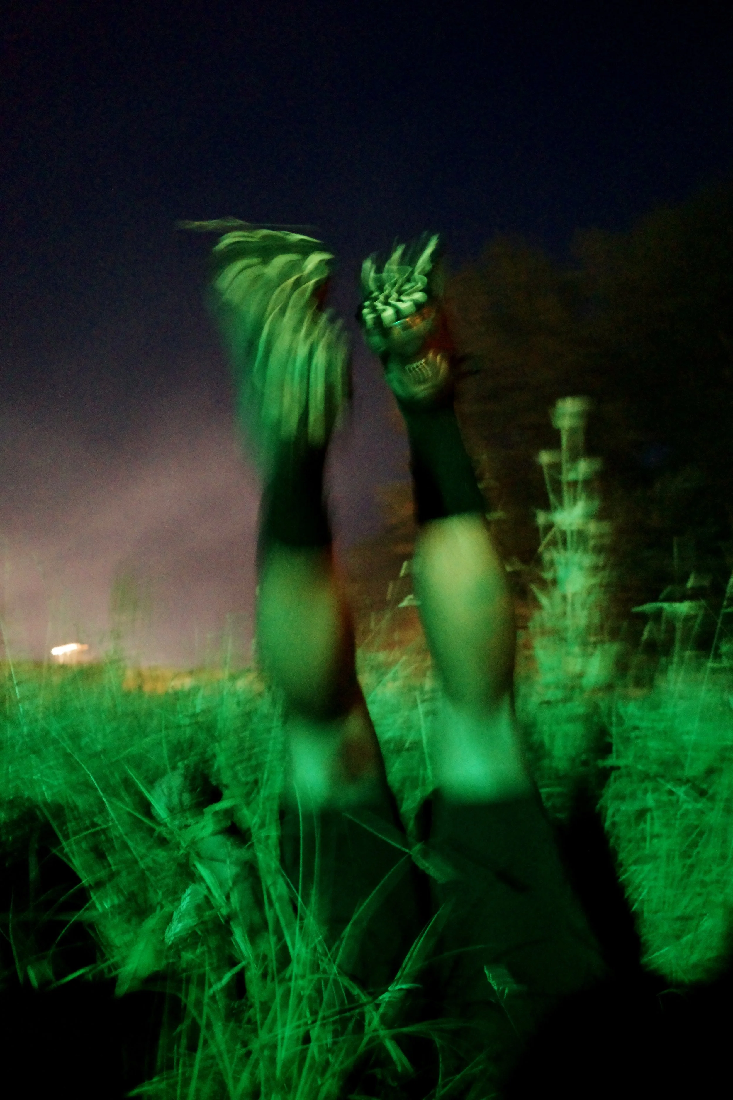 Hands with long nails illuminated by green lights in a grassy outdoor setting at night.