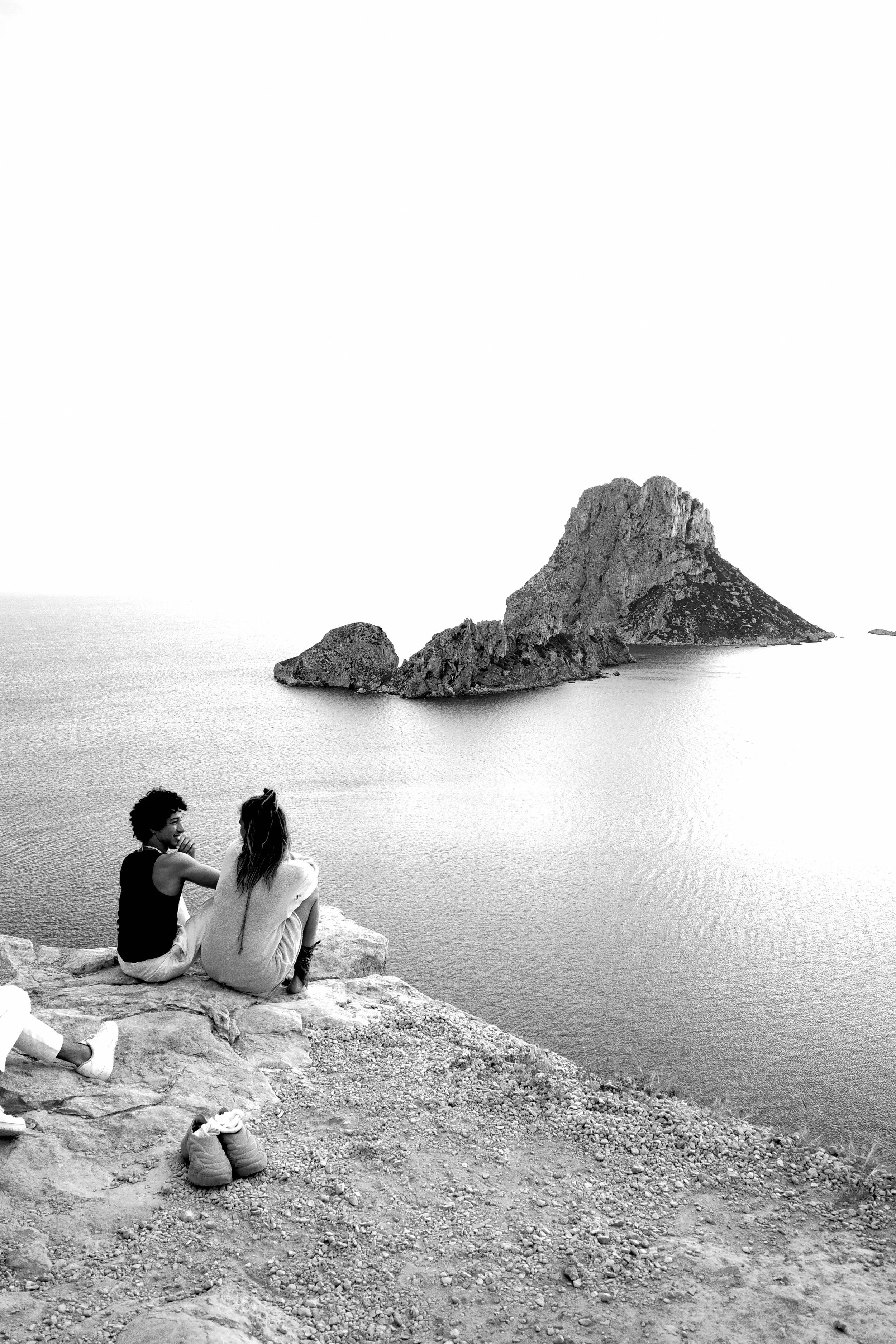 Two people sitting on a rocky shore overlooking the water, with a large rock formation island in the background.