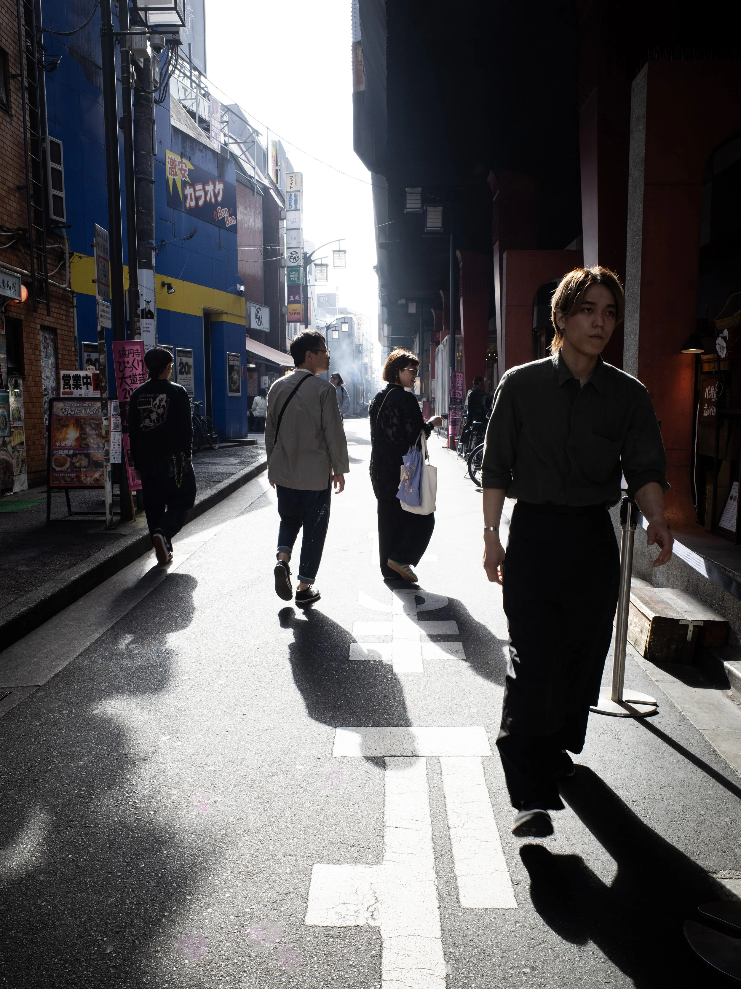 People walking on a city sidewalk in bright sunlight, with storefront signs and buildings in the background in Tokyo.