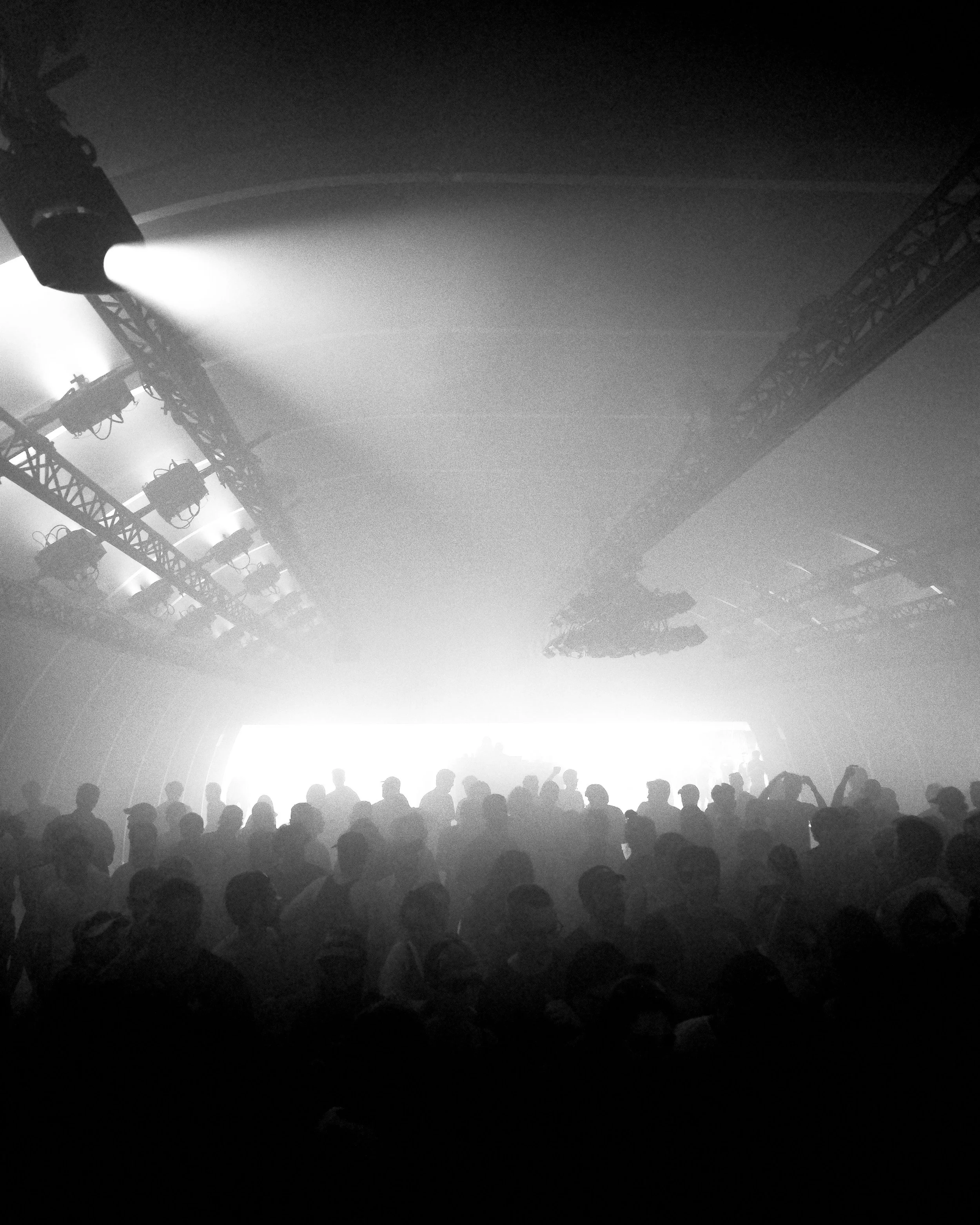 Black and white concert scene with a large crowd of silhouetted people facing a bright stage with multiple spotlights and lighting rigs at Dekmantel Festival.