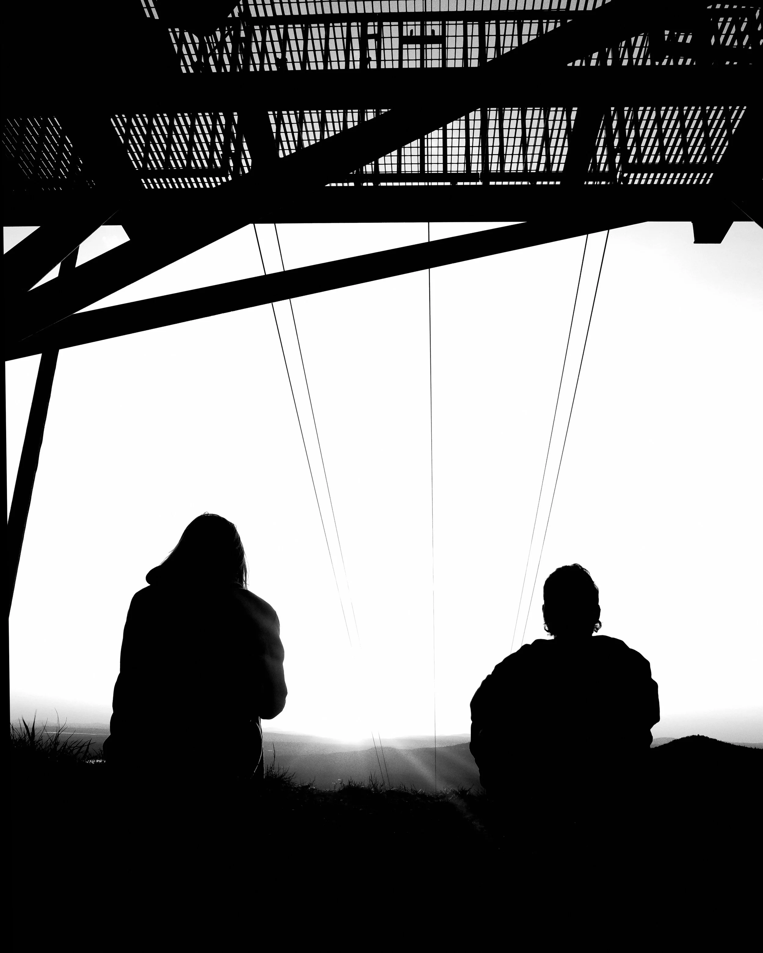 Silhouettes of two people sitting outdoors at sunset or sunrise, watching the horizon, with a power line and a large bridge structure overhead.