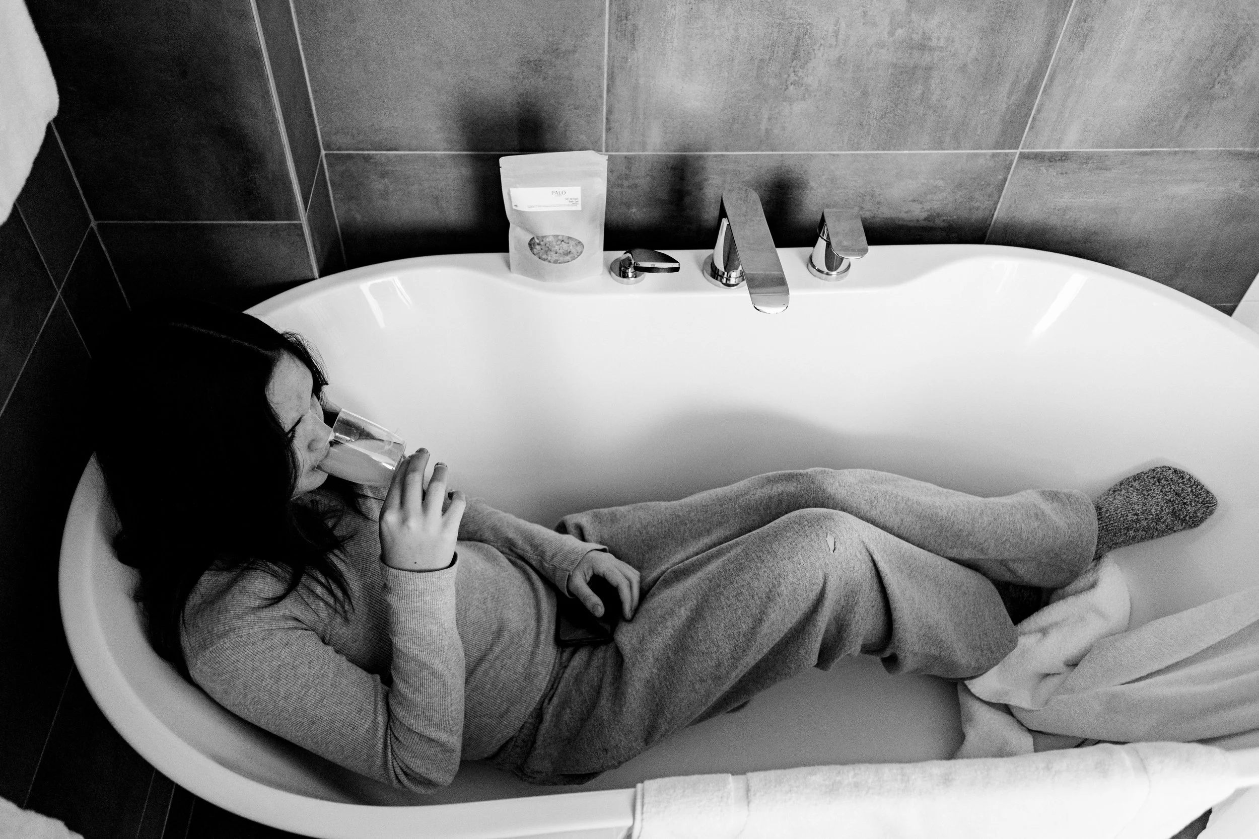 A dramatic black-and-white photograph captures a quiet, intimate moment of a bridesmaid during bridal prep. She is curled up inside a white porcelain bathtub wearing comfortable loungewear. She holds a smartphone in her hands without drinking from a 