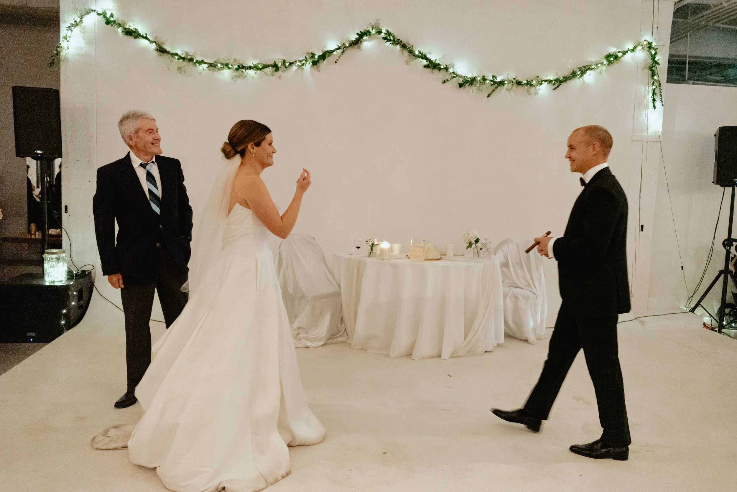 Bride beckons groom to her following father daughter dance. Father smiles to groom as the latter walks toward bride holding cigar