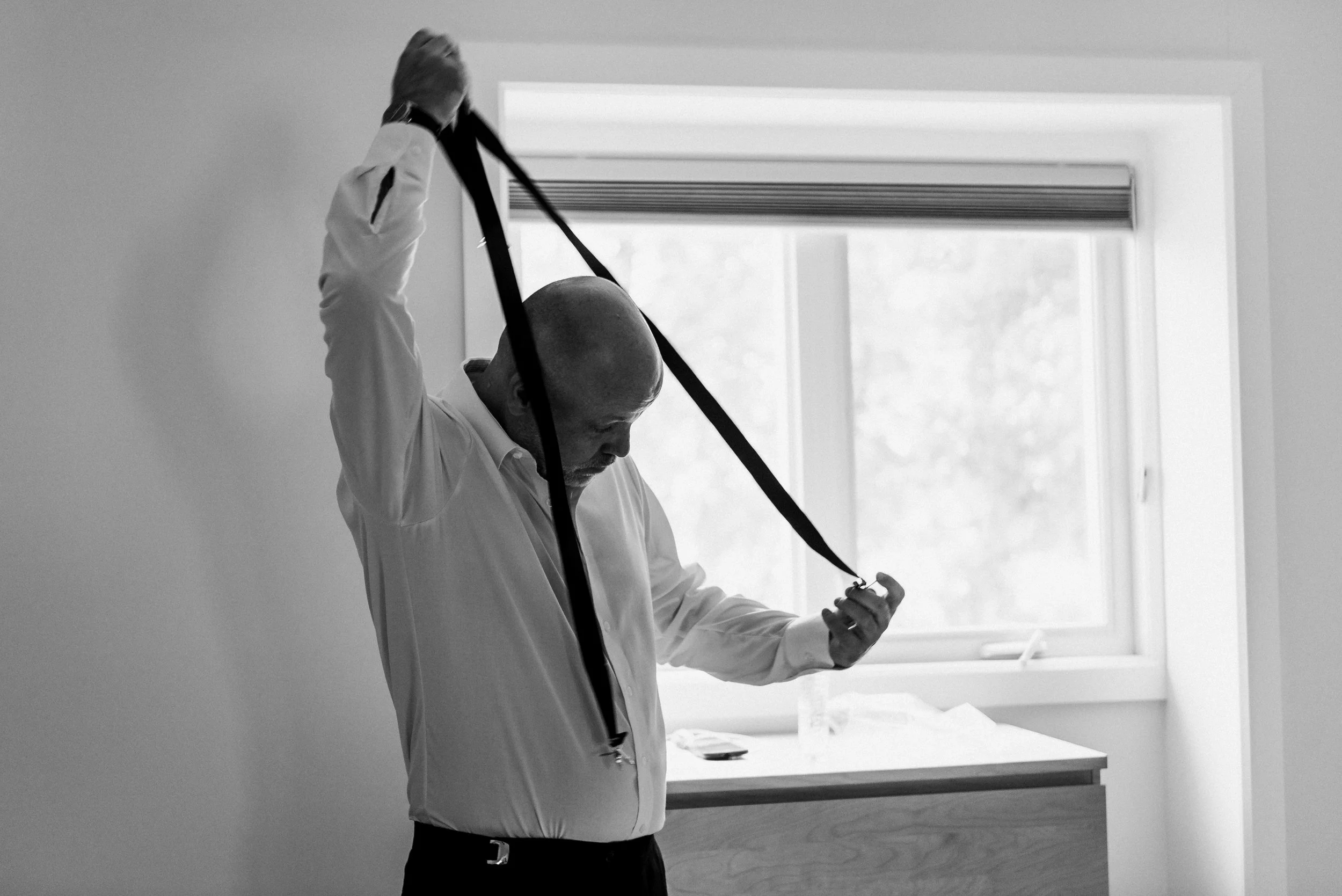 Black and white image of groom putting on his tie 