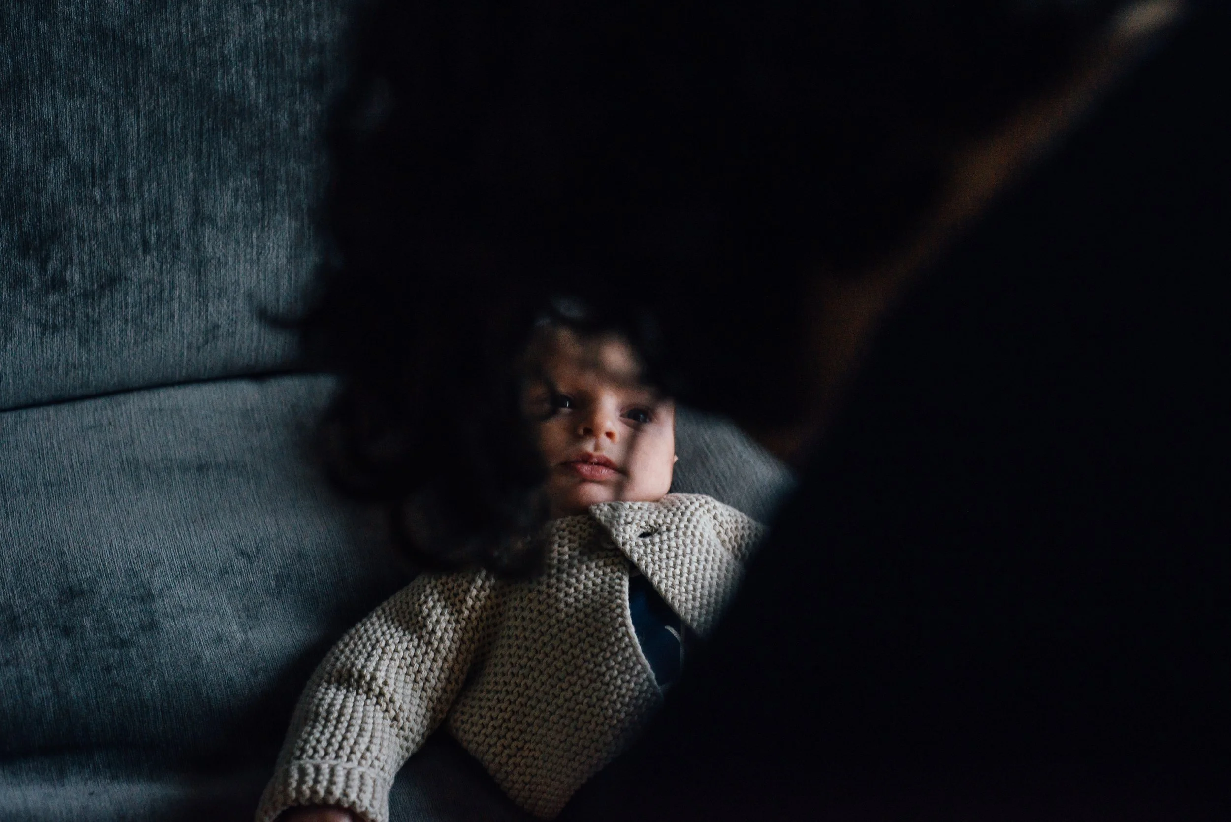 A young girl with curly hair lies on a dark sofa, looking upwards with her head resting on her hand, wearing a beige knitted sweater.